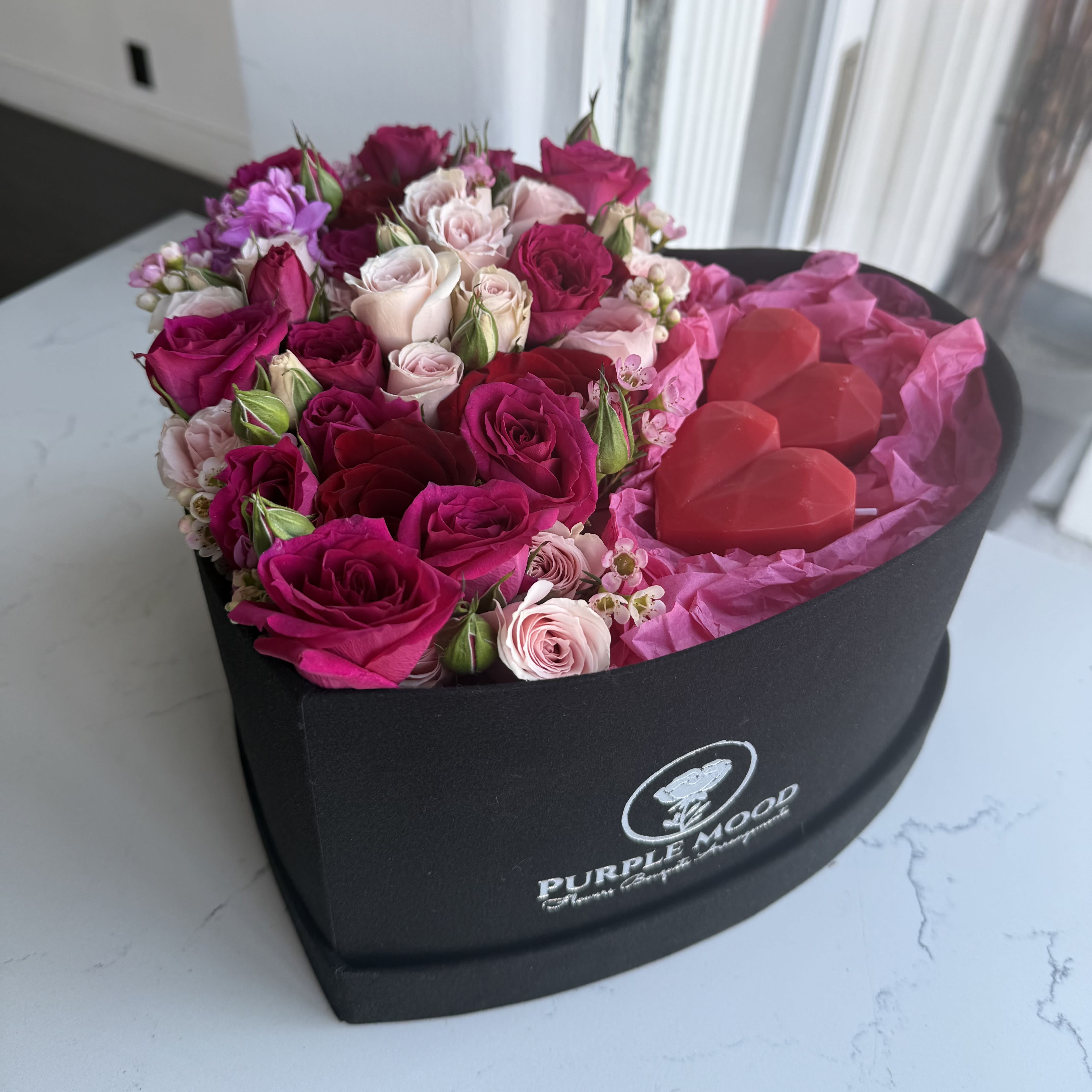 Heart box with candles -   Express your love with the elegant Heart Box with Candles, a stunning floral arrangement designed for romantic occasions. This heart-shaped box is filled with lush pink and red roses, soft blush spray roses, and delicate filler blooms, radiating beauty and charm. Accompanying the fresh flowers are handcrafted red heart-shaped candles, adding a unique and thoughtful element to this luxurious gift.  Perfect for Valentine’s Day, anniversaries, or any heartfelt celebration, the Heart Box with Candles will leave a lasting impression. 	•	Box Shape: Heart 	•	Includes: Fresh flowers and handcrafted heart-shaped candles 	•	Ideal For: Valentine’s Day, anniversaries, and romantic gestures 