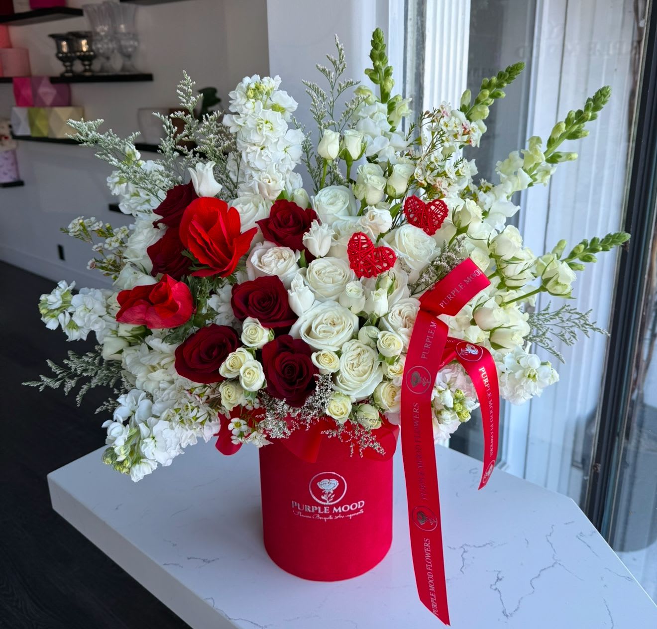 Love Love Box - The Love Love Box is a luxurious Valentine’s arrangement filled with radiant red and white roses, delicate white blooms, and lush greenery. Accentuated with romantic heart decorations and presented in an elegant red hatbox, this design symbolizes passion and pure affection. Tied with a signature Purple Mood ribbon, it’s the perfect gift to express your endless love.