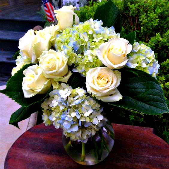 Blue Innocence  - Lovely design of mottled blue hydrangeas and creamy white roses with ferns designed in a clear glass vase. . NOTE: Blue and white hydrangeas may be used at certain times.   Blue Innocence is medium size of 3 designs. FAE-122