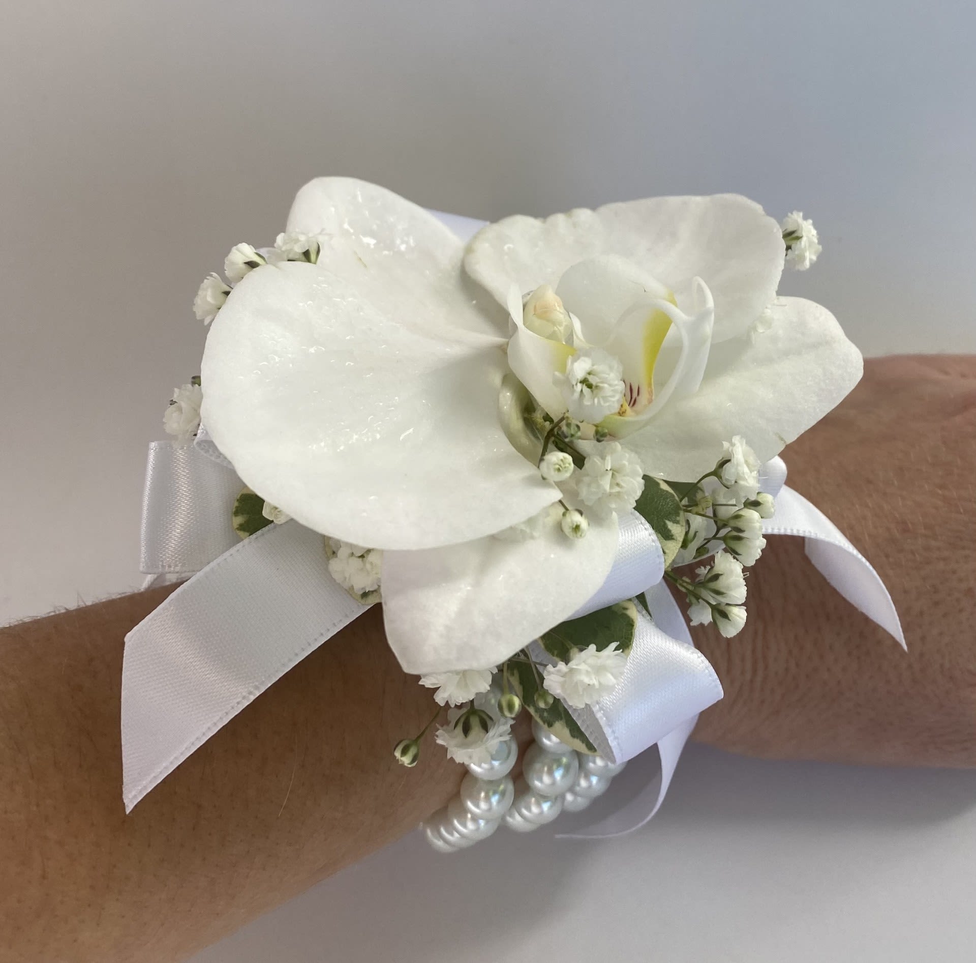 Corsage / Wrist - White Phalaenopsis - Orchid Wrist Corsage Elevate your formal attire with our Orchid Wrist Corsage, a stunning statement piece handcrafted to make your special night truly unforgettable. Designed with a pure white phalaenopsis orchid bloom and tailored to complement your unique style, this corsage is the perfect accessory for proms, homecoming dances, weddings, or any cherished celebration. Whether your vision is elegant, feminine, dramatic, or bold, our exquisite craftsmanship ensures a flawless design that reflects your aesthetic. -Key Features: **Fresh Large Orchid Bloom: Stately and elegant, a single orchid adds a delicate yet sophisticated charm. **Customizable Design: Include your dress color, preferred ribbon shade, and any floral requests to create a personalized piece. **Secure Bracelet Base: Comfortable, adjustable pearl band ensures a perfect fit for all-night wear. **Optional Trim Upgrades: Choose your favorite add-ins like rhinestones, pearls, or seasonal flowers for a bespoke design (quote provided upon request). To ensure your Orchid Wrist Corsage is a perfect match, please include the following in your order notes: Dress Color and Preferred Ribbon Color. Elevate your wrist corsage from a mere accessory to a bold statement. Order today and let us create a custom design that perfectly complements your style and makes your evening unforgettable!