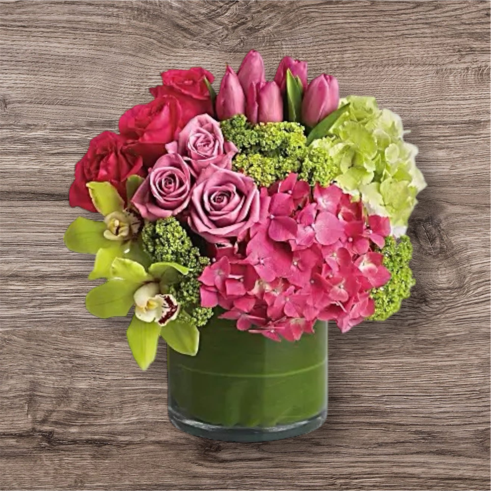  Sensations - A Touch of Tropical Serenity  Soft pinks and fresh greens come together in a lush, tropical-inspired arrangement that brings harmony and a calming energy to any space. Designed to uplift the senses and refresh the soul with a perfectly balanced palette of nature’s most serene hues.  • Features green and pink hydrangeas, green cymbidium orchids, hot pink and lavender roses, pink tulips, and green viburnum.  • Artfully arranged in a clear glass cylinder vase lined with tropical green ti leaves.  • All-around design for a beautiful view from every angle.   