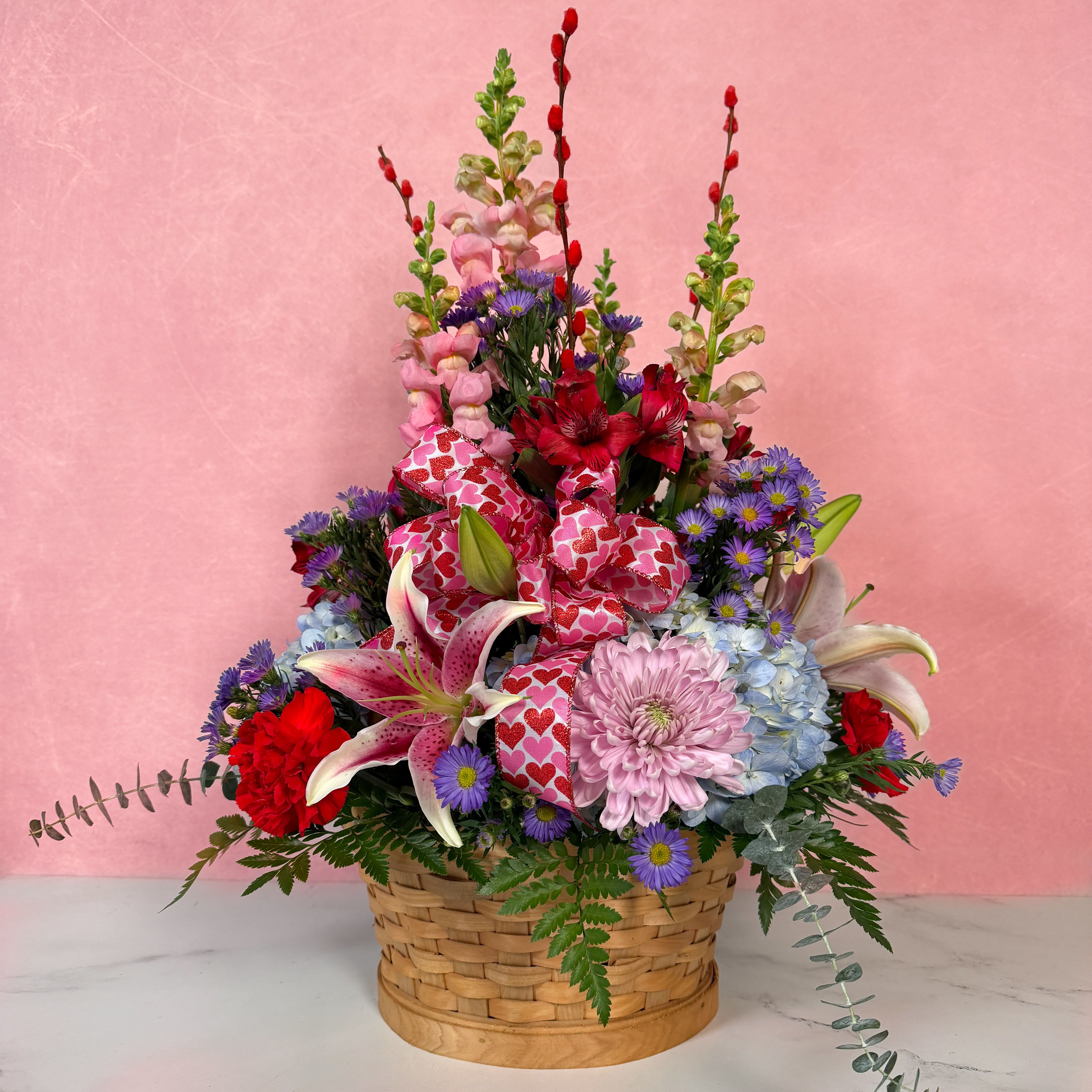 Basket of Love - Our Basket of Love is a grand display of affection, with a variety of colorful blooms, including blue hydrangea, stargazer lilies, pink snapdragons, and more! Featuring a Valentine's heart bow to tie it all together, this basket arrangement is the perfect way to show someone that you love them this Valentine's Day!  Approx. L 22” x H 24”