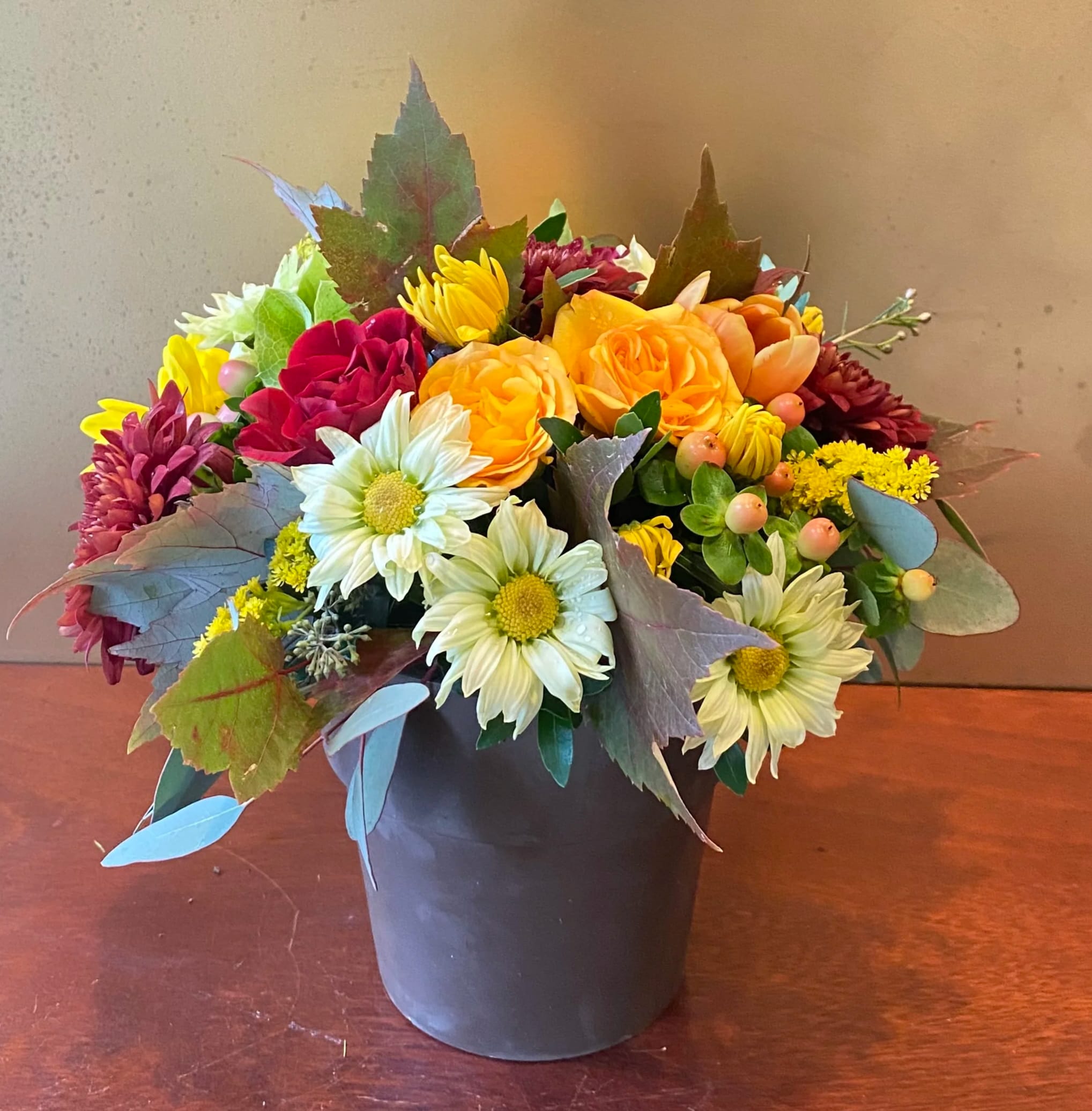 Sugar Maples - Chrysanthemums in autumnal colors of yellow, bronze and green - set off by fiery orange roses, seeded eucalyptus and hypericum - nestle together with brilliant fall leaves to create a bright floral arrangement that's reminiscent of a walk in the woods at sugar maple time.  Orange spray roses, yellow viking spray chrysanthemums, bronze cushion spray chrysanthemums, green button spray chrysanthemums, red hypericum, seeded eucalyptus, fall leaves and salal are arranged in a terra-cotta pot.