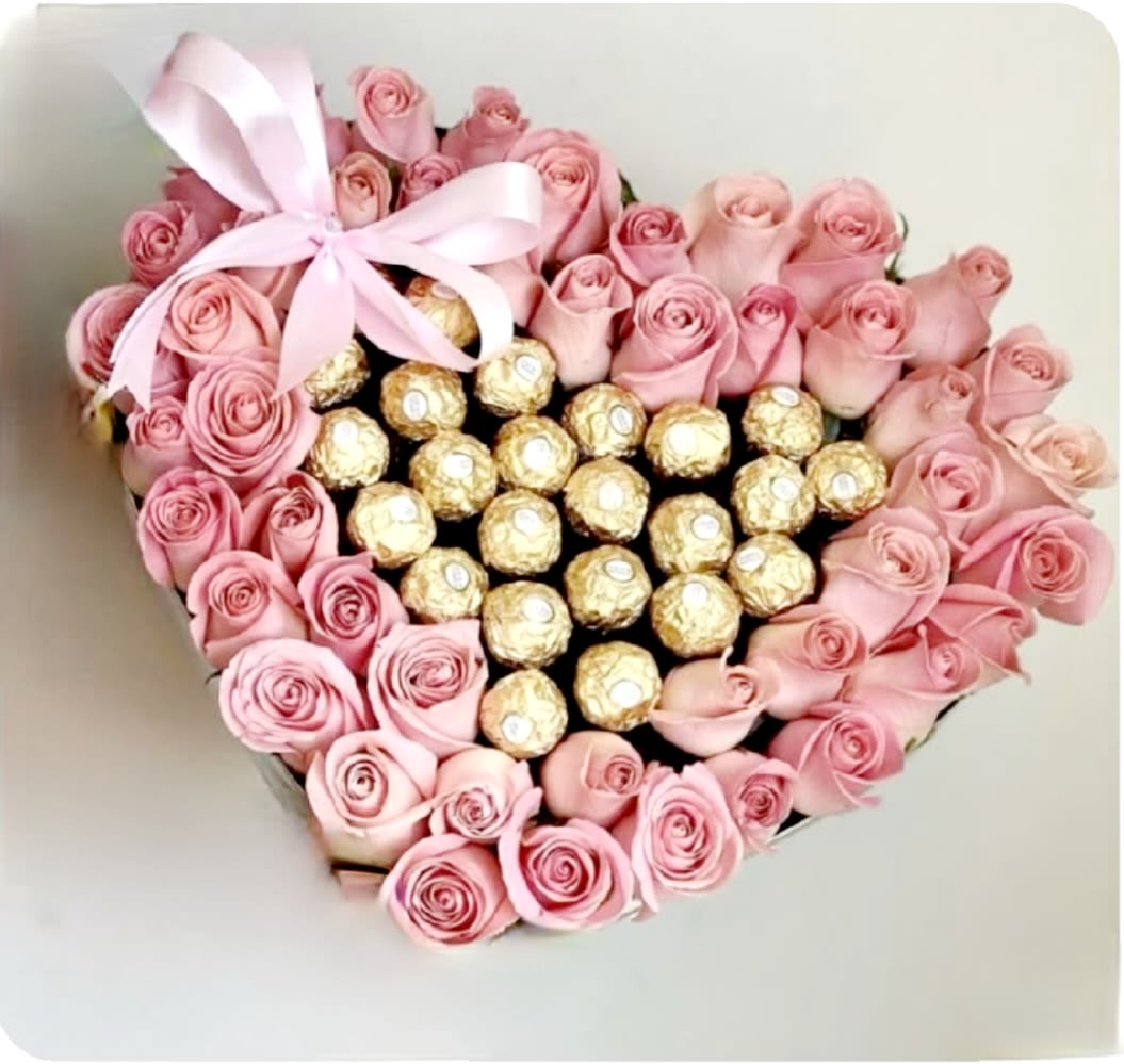 Pink and Gold Gems by Pretty in Petals  - This heart-shaped arrangement features a border of neatly arranged pink roses encircling a central cluster of Ferrero Rocher chocolates. A pink ribbon bow is placed at the top left corner, adding a decorative accent. The composition blends floral elegance with gourmet appeal, making it ideal for a romantic or celebratory gift.  Description: One size only premium - 48 Pink, Red, White or Mixed Roses - with a custom Handwritten Valentine Card   Flowers: • 	Pink Roses: These form the entire outer border of the heart. They are tightly packed and uniformly colored, likely standard hybrid tea roses or spray roses. Their soft pink hue suggests themes of admiration, sweetness, and affection. Candy: • 	Ferrero Rocher Chocolates: The center is filled with neatly arranged Ferrero Rocher chocolates, known for their gold foil wrapping and round shape. They are placed in a grid-like pattern, creating a rich, indulgent focal point.  Changes may be made due to product inventory, but will honor the inspirational photo as closely as possible.