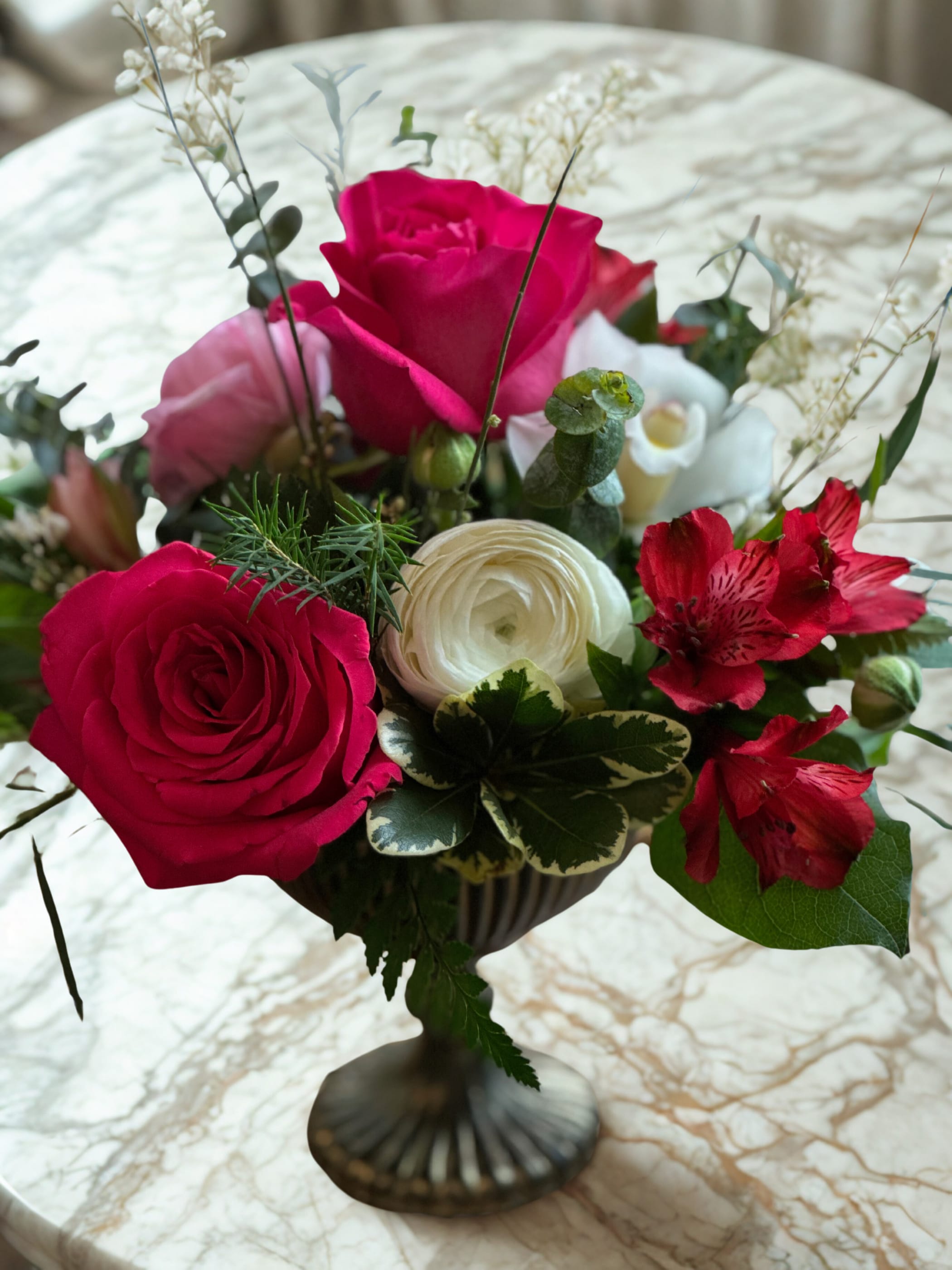 Compote Glass Container - Designed in a classic footed glass bowl, this compote flower arrangement offers a romantic, loose, garden-style aesthetic. Lush greenery and a thoughtfully curated mix of blooms gently spill over the edges, creating an airy, asymmetrical design. We begin by layering fresh foliage, then add roses alongside other focal flowers and soft fillers to achieve a whimsical, natural feel. This popular arrangement style is perfect for any occasion and is available in a variety of beautiful color palettes. 