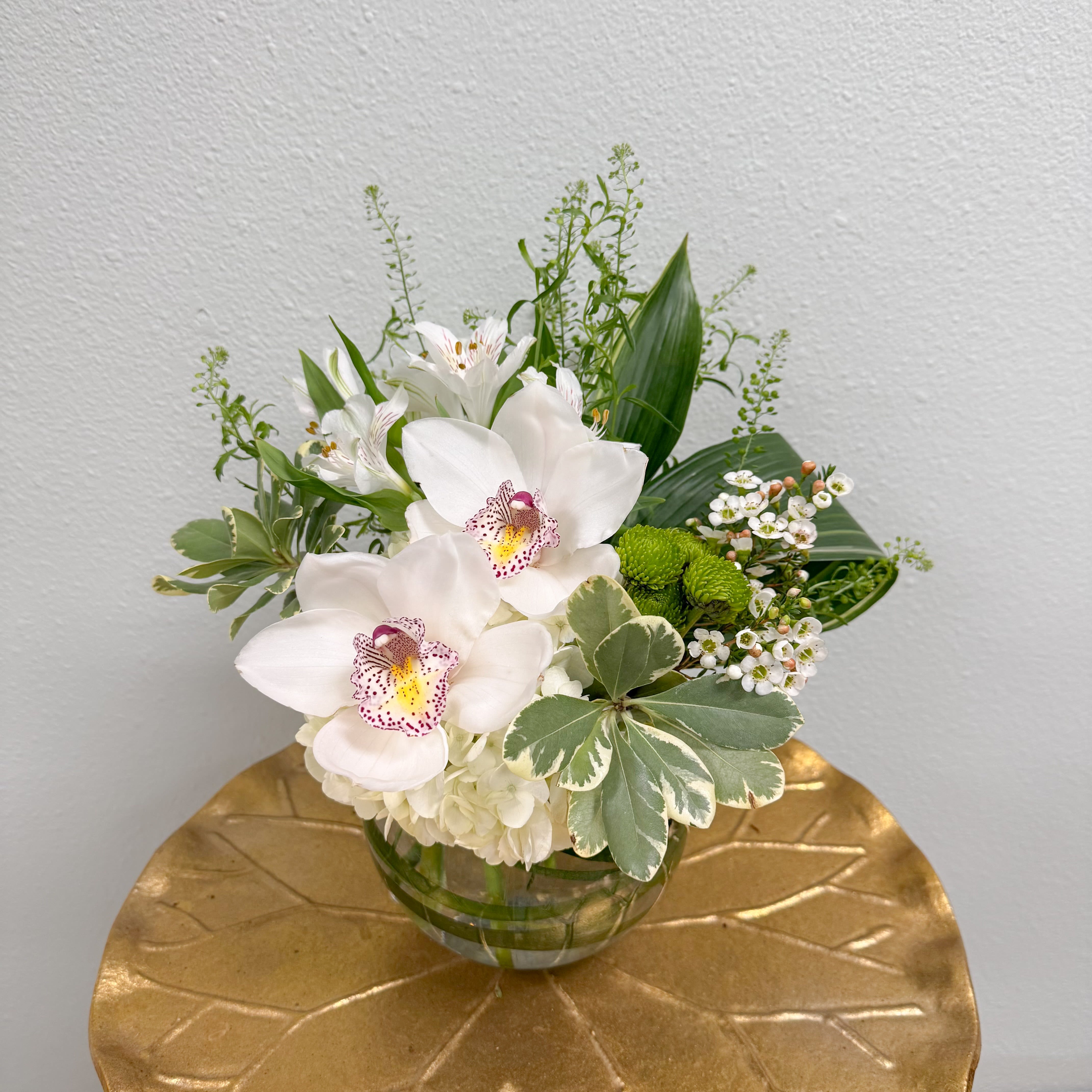 Calming Tropics - Cymbidium orchids are the star of this elegant arrangement complimented by fresh green and white blooms and tropical leaves in a bubble bowl. This one-sided arrangement is perfect for a sweet gesture or small places.