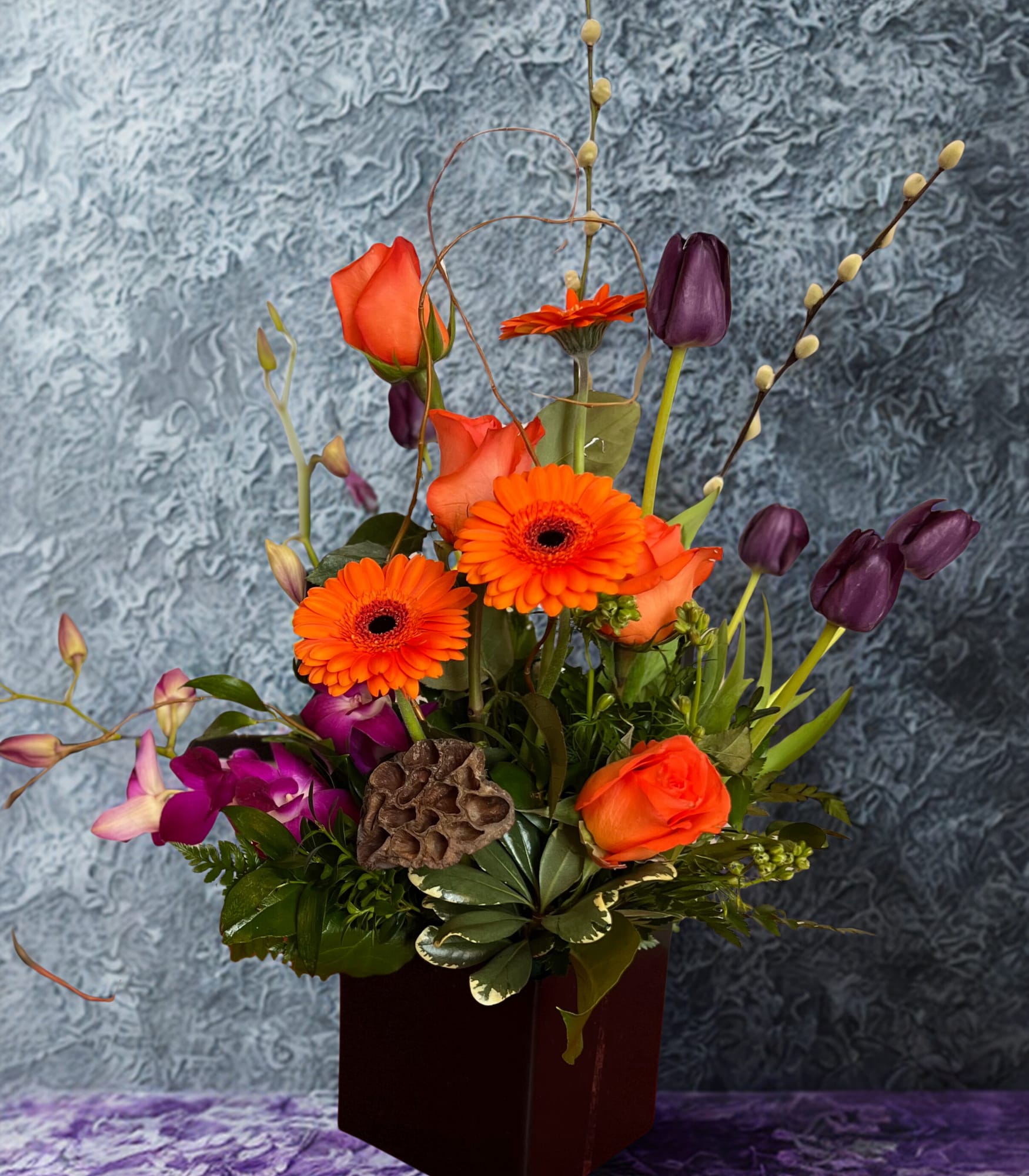 A Composition Creation - This striking arrangement features a vibrant blend of orange gerbera daisies, purple tulips, orange roses, and graceful dendrobium orchids. Thoughtfully designed in a sleek black ceramic cube, the focus is placed on the artistic composition of the blooms themselves. The result is a bold yet elegant floral design with a clean, modern feel—perfect for making a contemporary statement. 