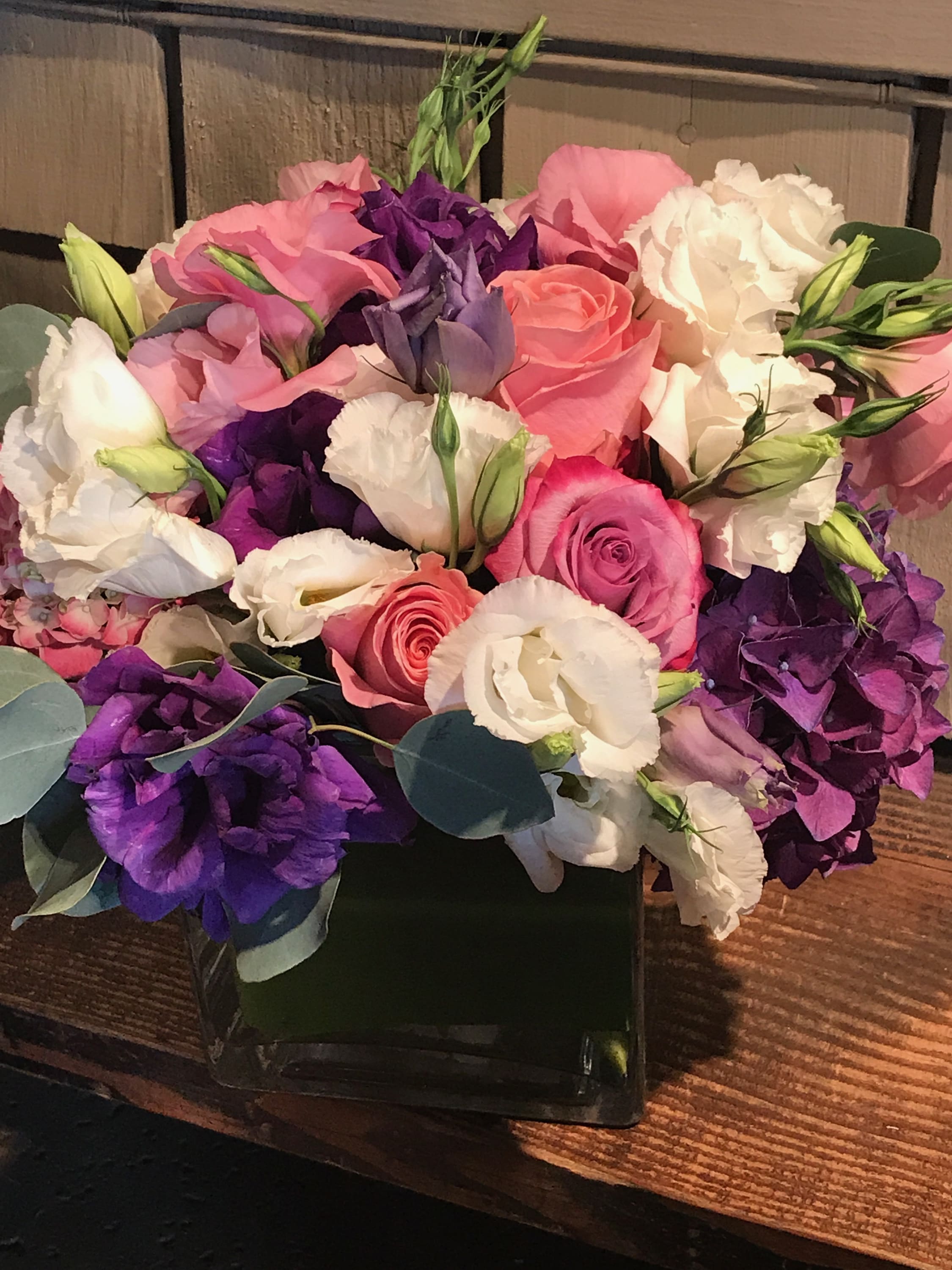 Castera Rose - Purple, pink hydrangeas mixed with purple, pink, white lisianthus and castera roses wrapped in tea leaves arranged in square glass vase.