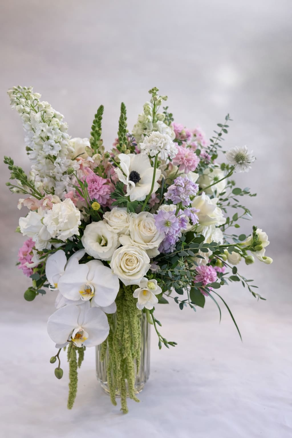Pastel Spring - Flowers: roses, stock, delphinium, orchids, snapdragons, lisianthus, anemones, dahlias, freesia, eucalyptus, and amaranthus. Vase: Tall lined vase approximately 6x8
