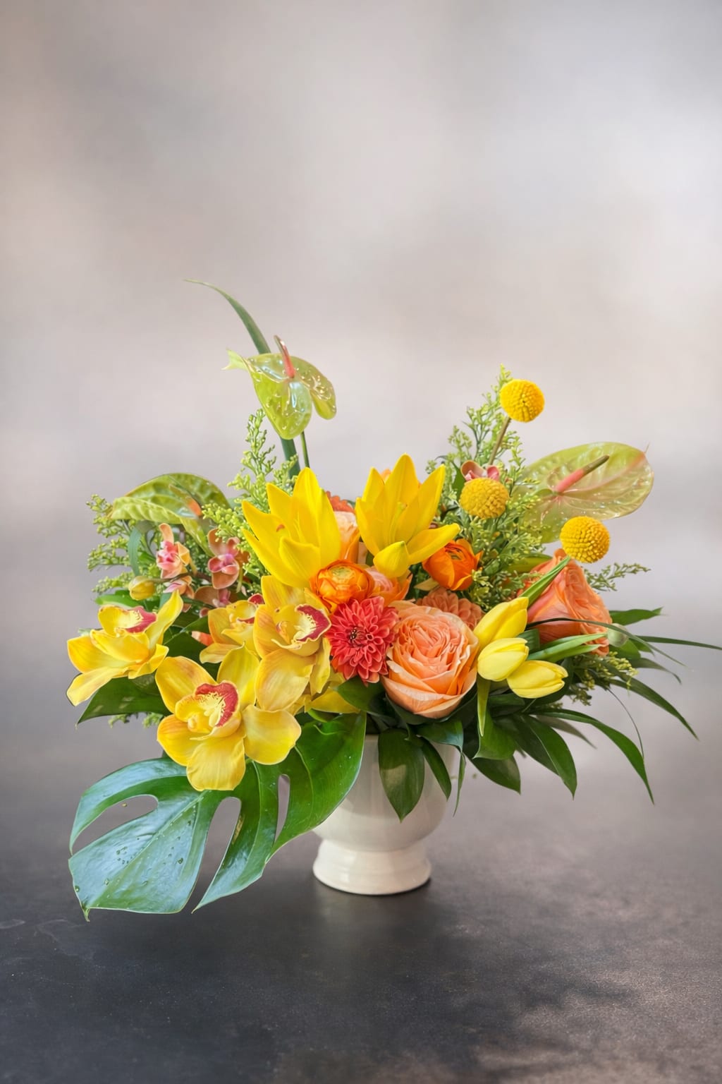 Delight Me - Flowers: yellow cymbidium orchids, yellow asiatic lilies, billy balls, anthurium, kahala roses, tulips, solid aster, monstera leaves, and greenery Vase: small ceramic white compote