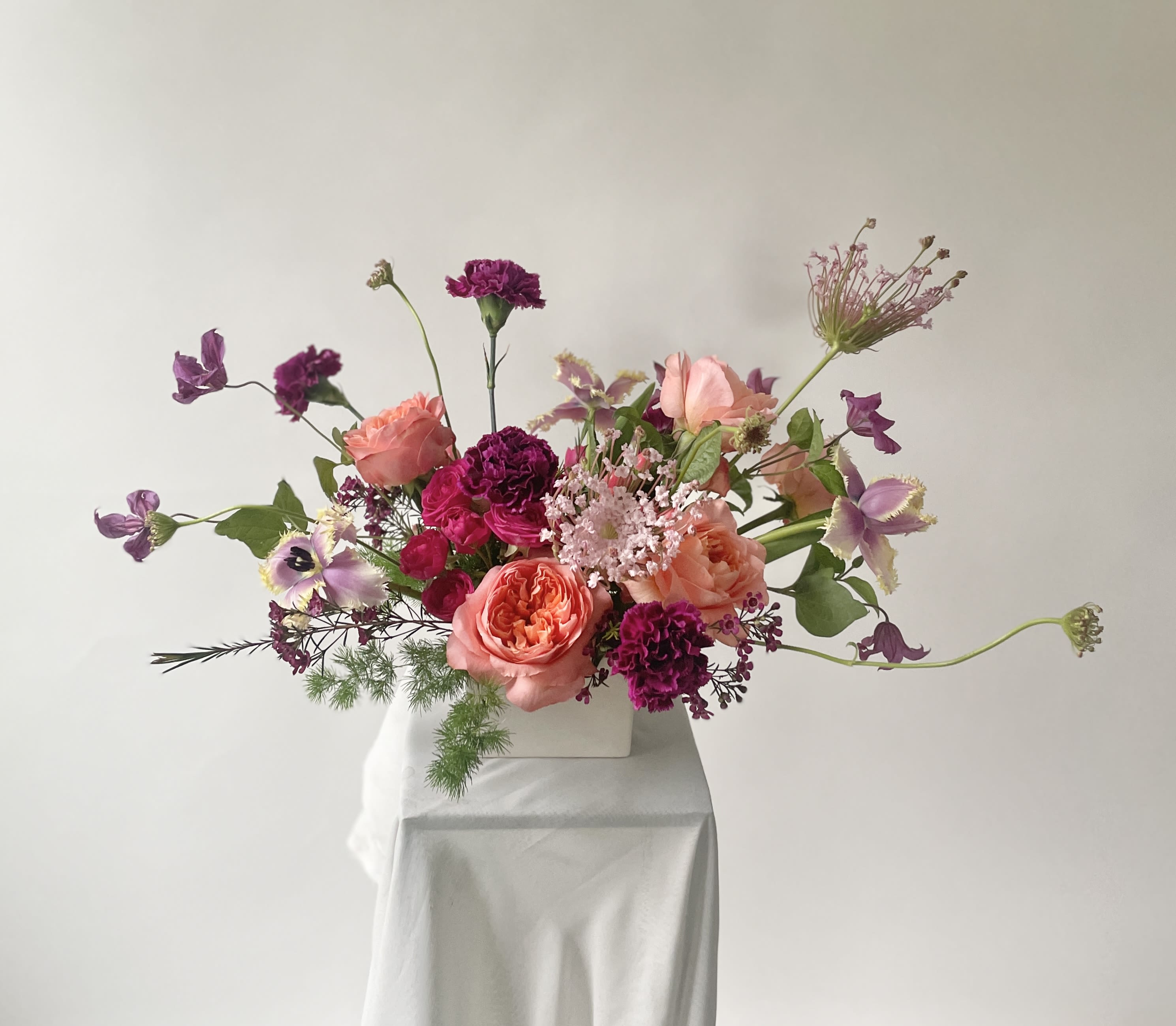 Berry Blossom - Vibrant, romantic, and full of personality, Berry Blossom is a floral arrangement that delights the senses with its lush textures and joyful palette. Featuring a striking mix of berry-toned blooms, coral garden roses, and delicate accents that dance with movement, this design blends the untamed beauty of a blooming meadow with a modern, sculptural touch.  Crafted in our signature garden-forward style, and in a clean, modern vessel, Berry Blossom is designed to make a bold impression—from dining tables to entryways and every special corner in between.