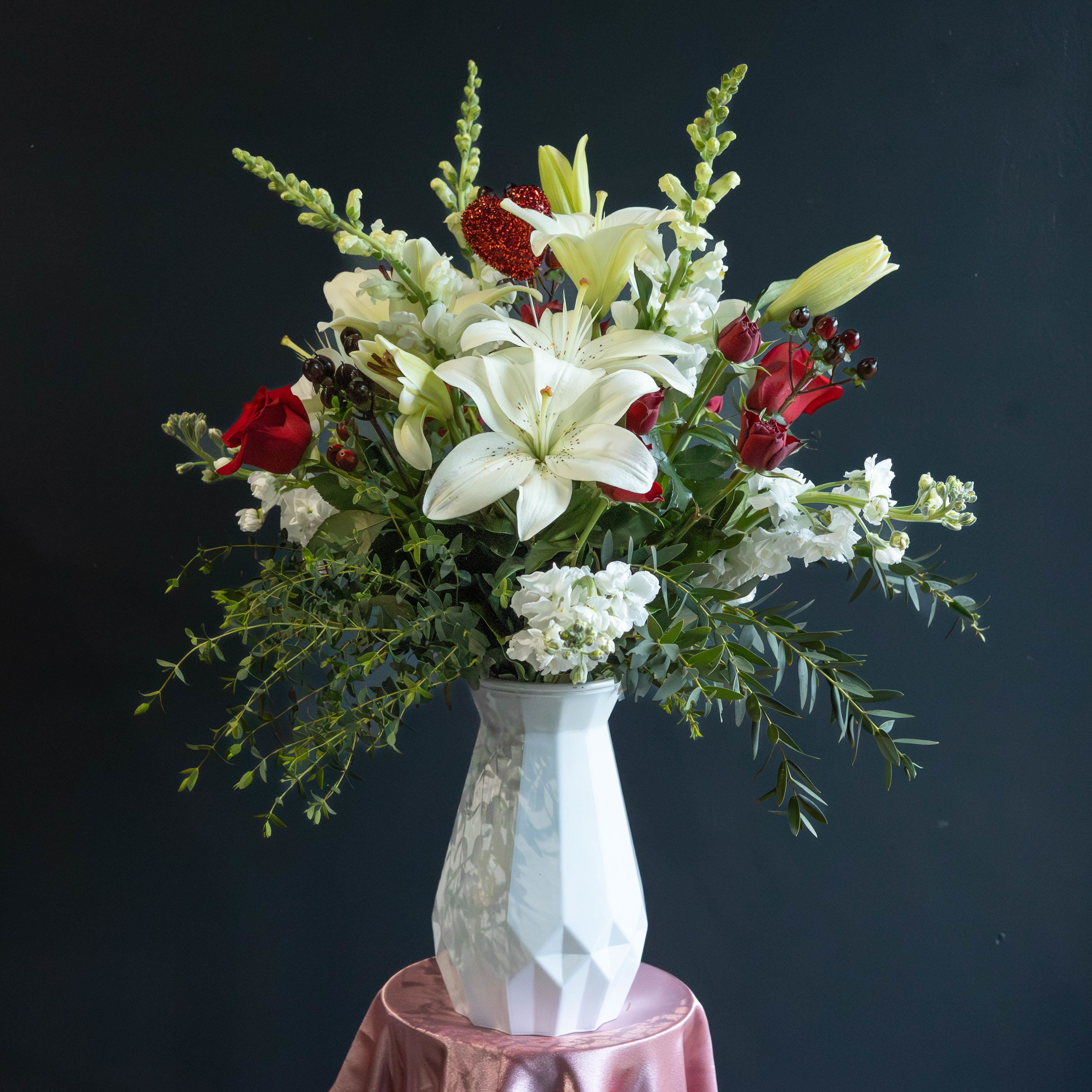 Love Story - Love Story is an elegant Valentine’s Day arrangement that tells a love story at first glance. Featuring graceful white lilies at the center, surrounded by classic red roses, soft white blooms, and lush greenery, this design blends romance with timeless beauty. A subtle heart accent adds a sweet Valentine’s touch, while the modern white faceted vase completes the look with clean sophistication. Designed to feel both heartfelt and refined, Happily Ever After is a stunning way to celebrate love, commitment, and meaningful moments.