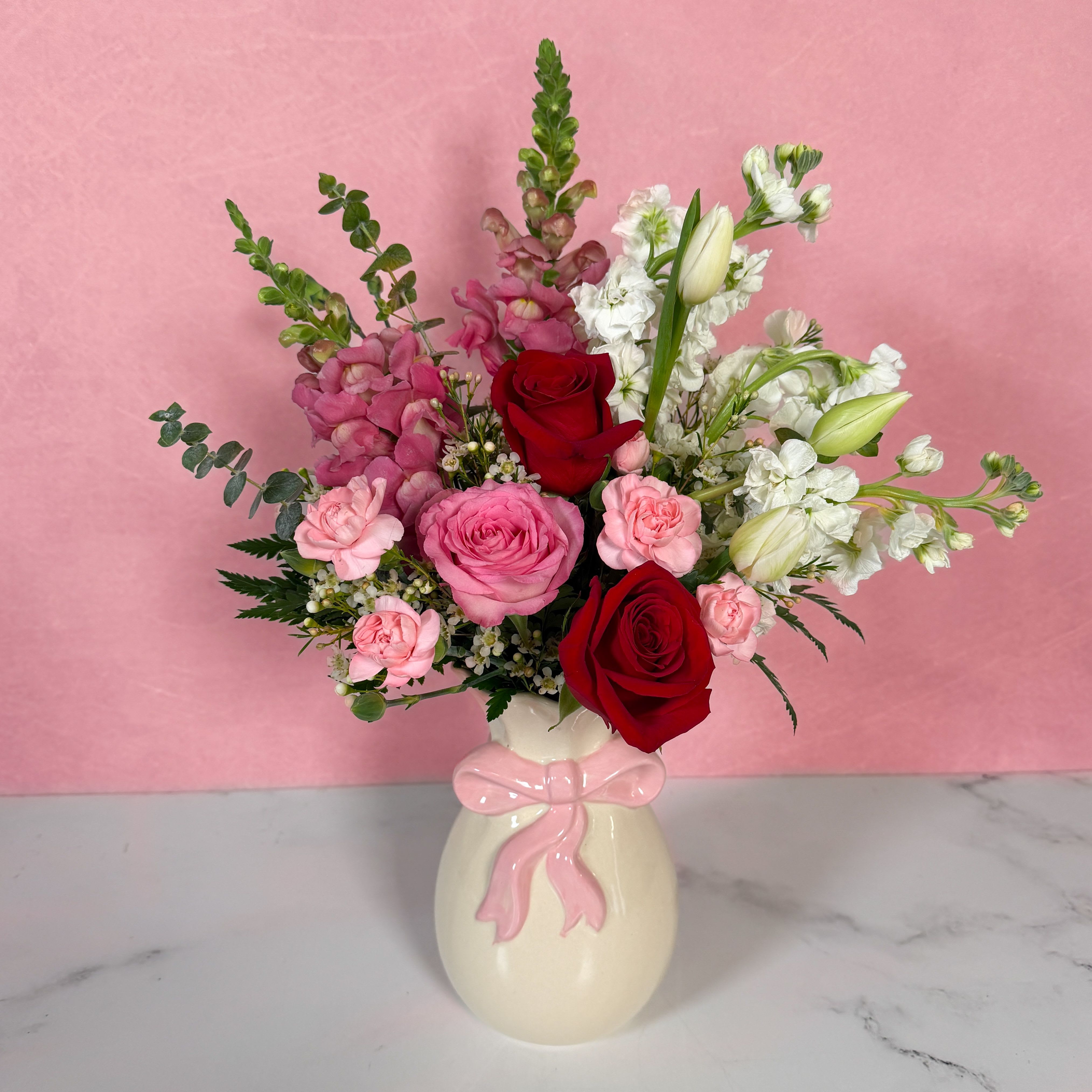 Tied With Love Bouquet - Everything you could want for Valentine's Day, all tied up in a nice little bow! Our Tied With Love Bouquet is a delightful display of Valentine's blooms, including lovely red and pink roses, pink snapdragons, delicate white tulips, and more! These dazzling flowers are presented in an adorable vase with a pink bow design "wrapped" around the neck of the vase! If you want a stylish and fun gift for your sweetheart this Valentine's Day, our Tied With Love Bouquet is sure to be a hit!  Approx. L 15" x H 19"
