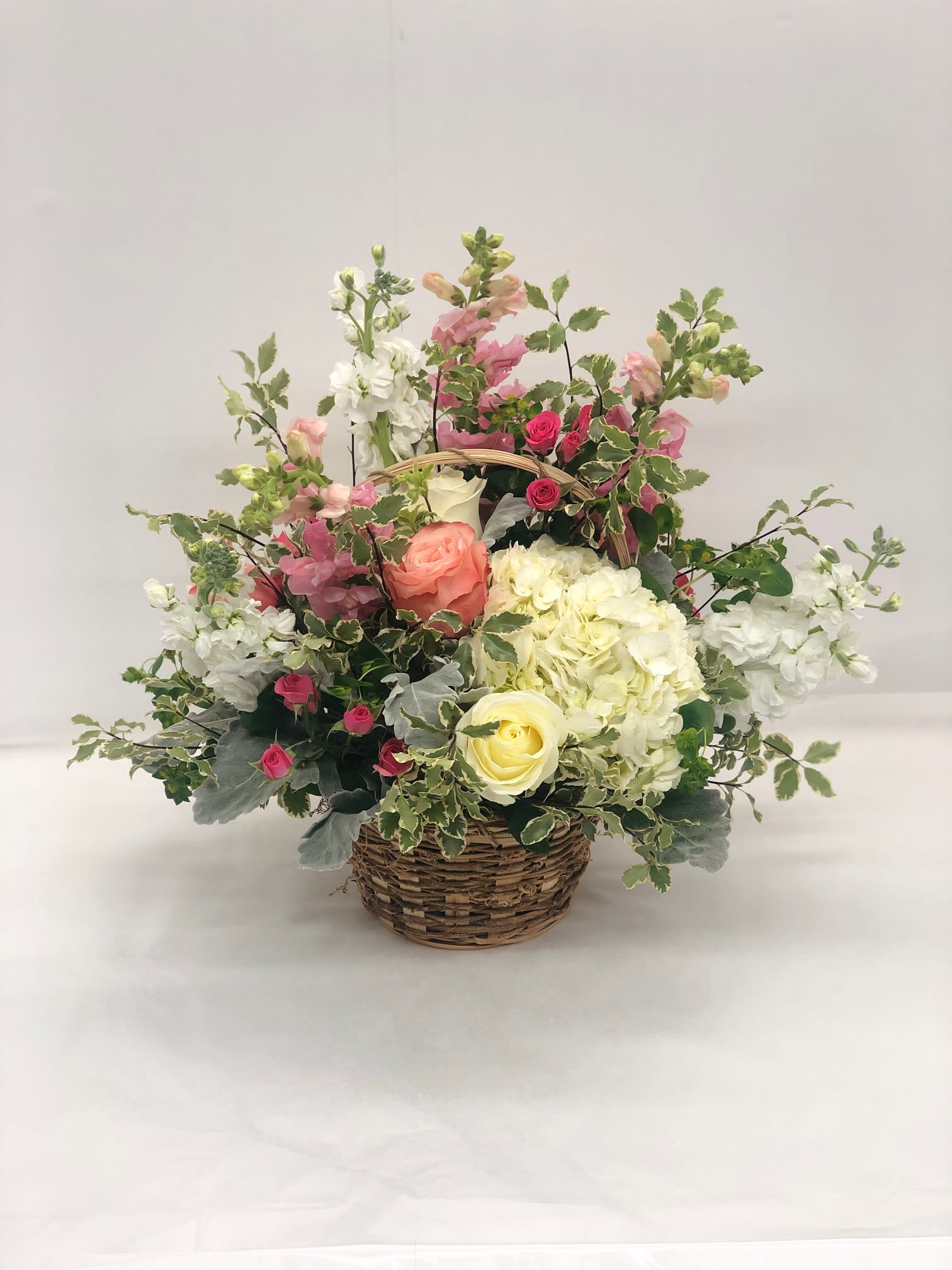 Designer's Bountiful Garden - White and salmon roses made the eyes dance while surrounded by pink larkspur, cream gilly flower, peach spray roses, clouds of white hydrangea, dusty miller stems, and lush greens, arranged to perfection in an oval stained woodchip basket that helps to blend soft sophistication with raw, rustic appeal. That perfect happy birthday, thinking of you, or thank you gift!   Approximately 16" tall x 11" wide   