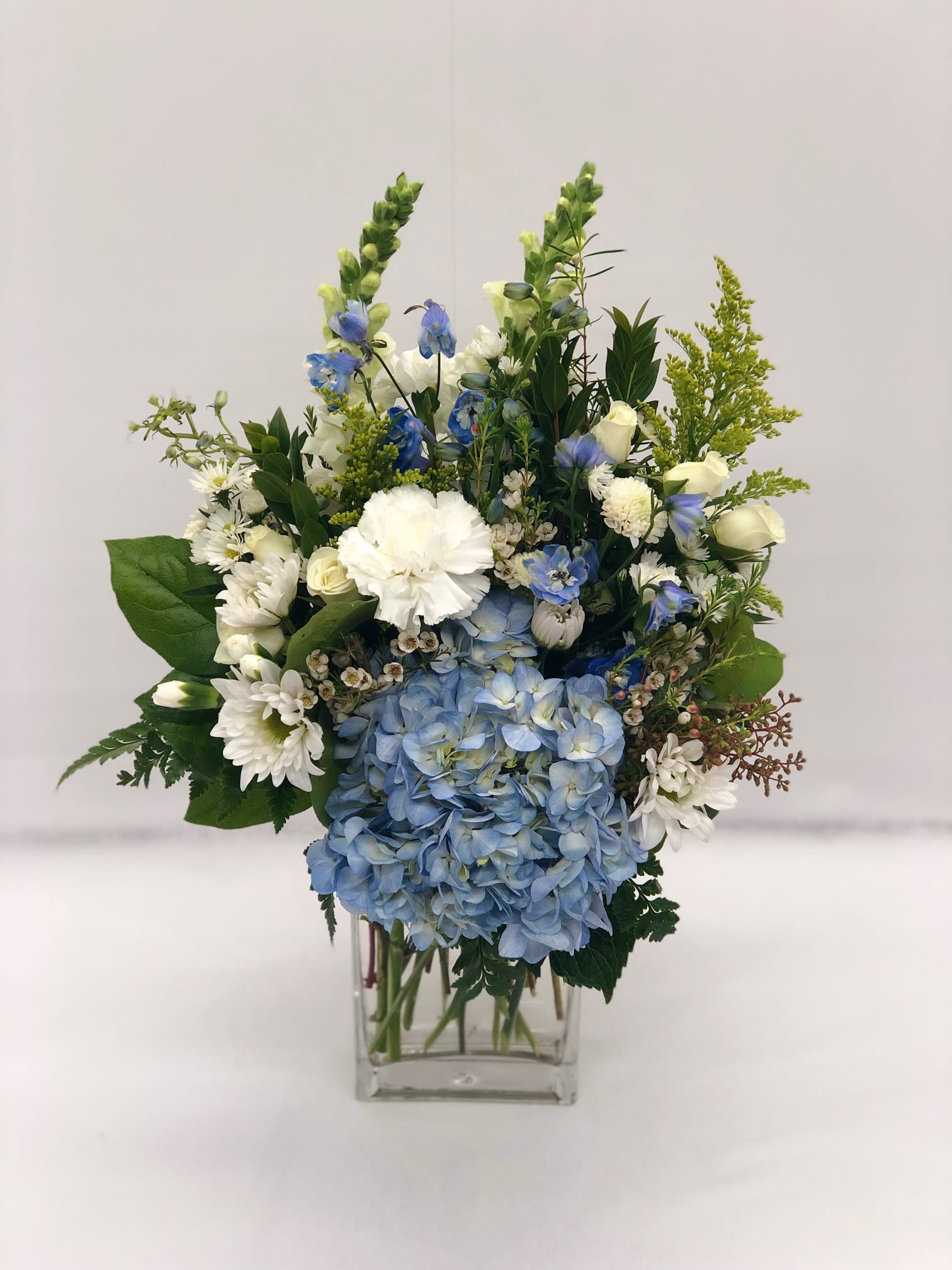 Blue Skies - Beautiful assortment of blue and white flowers arranged together in a short rectangular clear vase. 