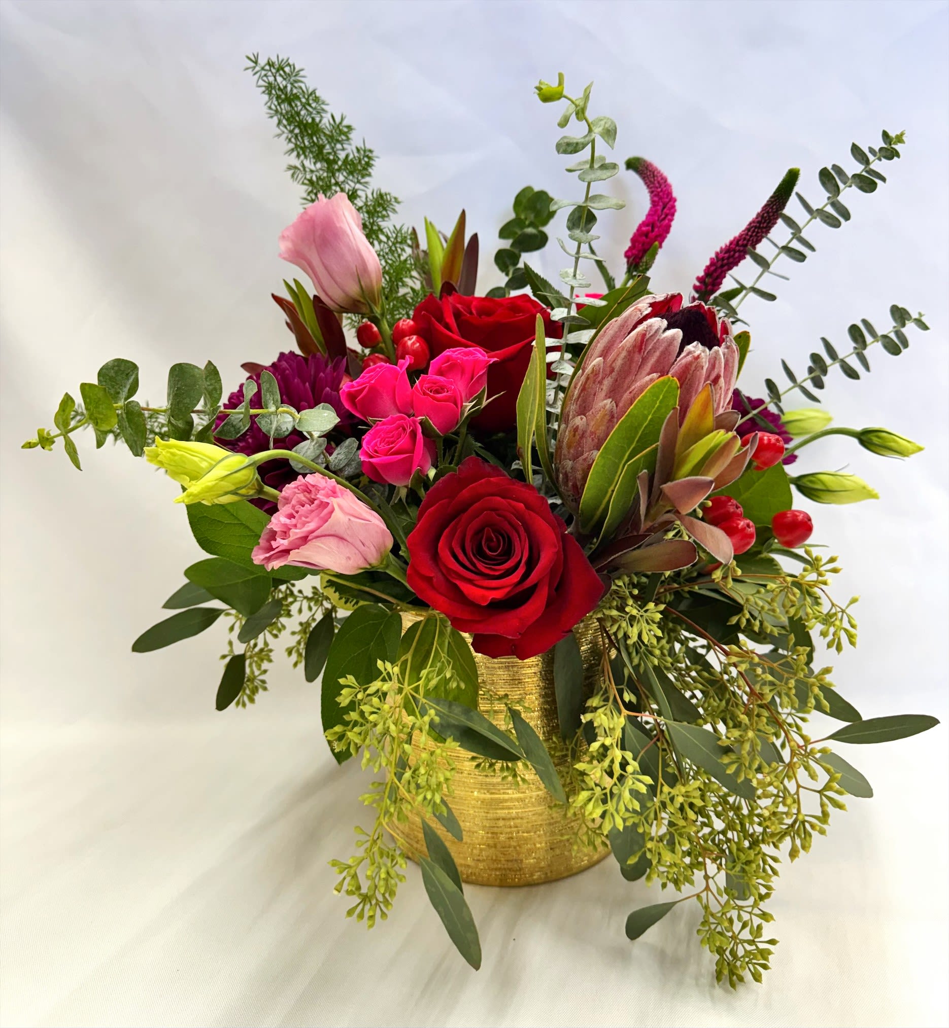 Hottie - Hottie Bouquet  A sultry explosion of passion and color. Featuring lush red roses, hot pink spray roses, and soft light pink lisianthus, this arrangement is elevated with striking mauve protea, rich purple chrysanthemums, and juicy red hypericum berries. Seeded eucalyptus and layered greenery add texture and balance, creating a look that’s equal parts romantic and unapologetically bold. Designed to turn heads and steal hearts, HOTTIE is made for love that burns bright.  