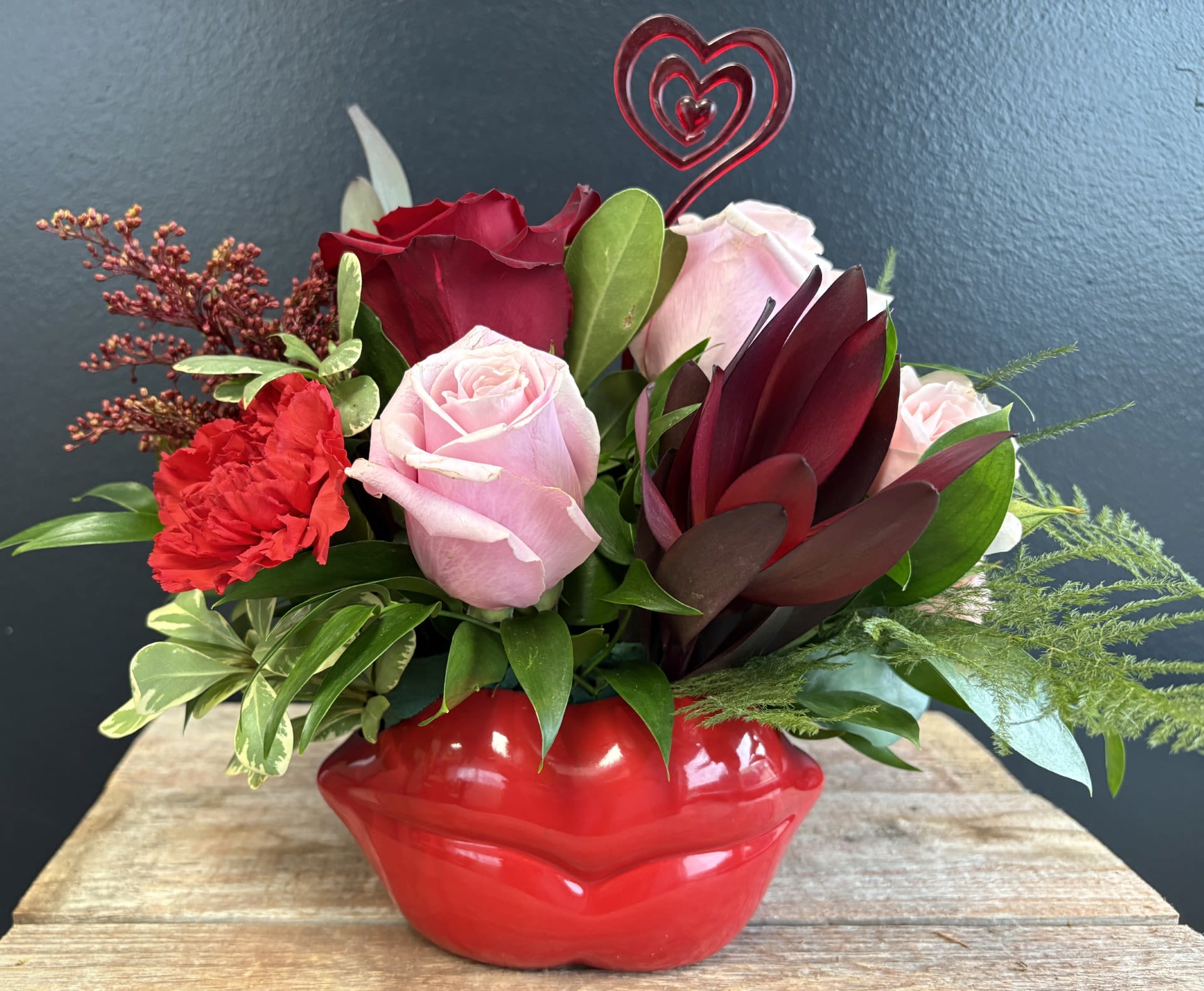 Hot Lips  - A striking lip‑shaped container becomes the heart of this Valentine’s design, its smooth curves echoing the sweetness of a kiss. Nestled within the bold silhouette, lush blooms in shades of ruby red, blush pink, Colors may vary in the shades or pinks etc.
