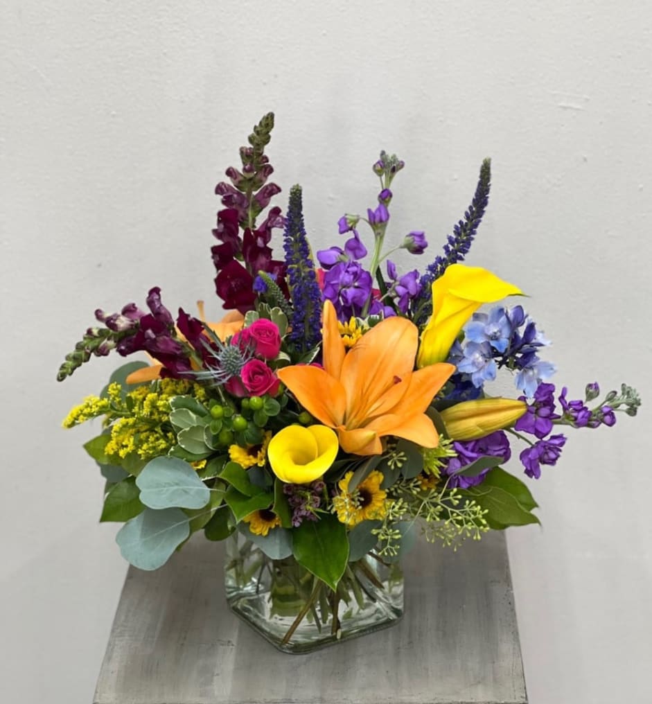 Prism - Assortment of lilies, calla lilies, stock, snapdragons, spray roses, delph., veronica, thistle, viking mums, hypercium berries in a SHORT clear cube vase.  Flowers and colors may vary depending on availability.