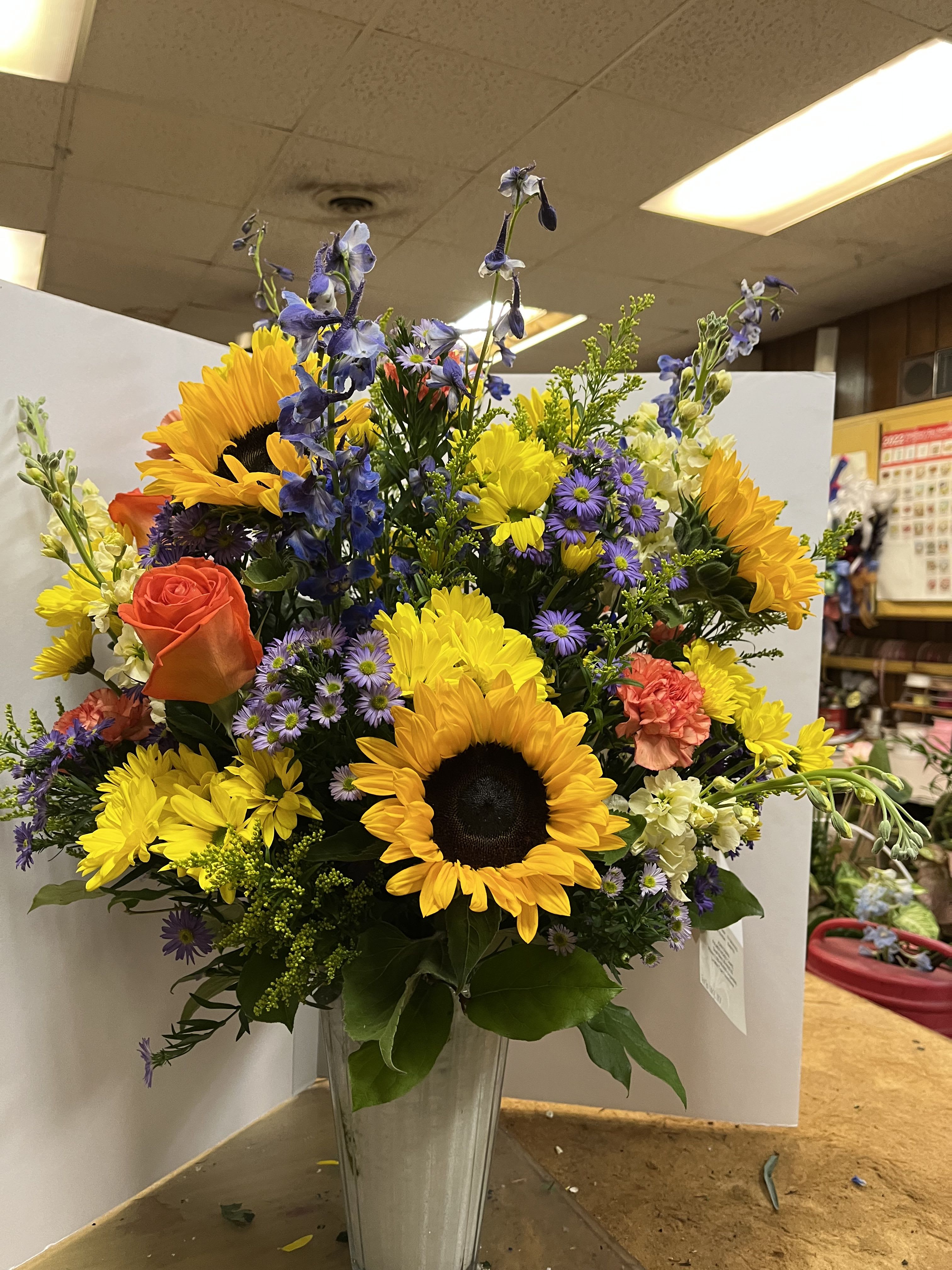 WS-0804 Linda - Vased arrangement including sunflowers, orange roses, orange carnations, blue delphinium, yellow daisies,  purple monte casino, yellow snap dragons, solidago and salal.  