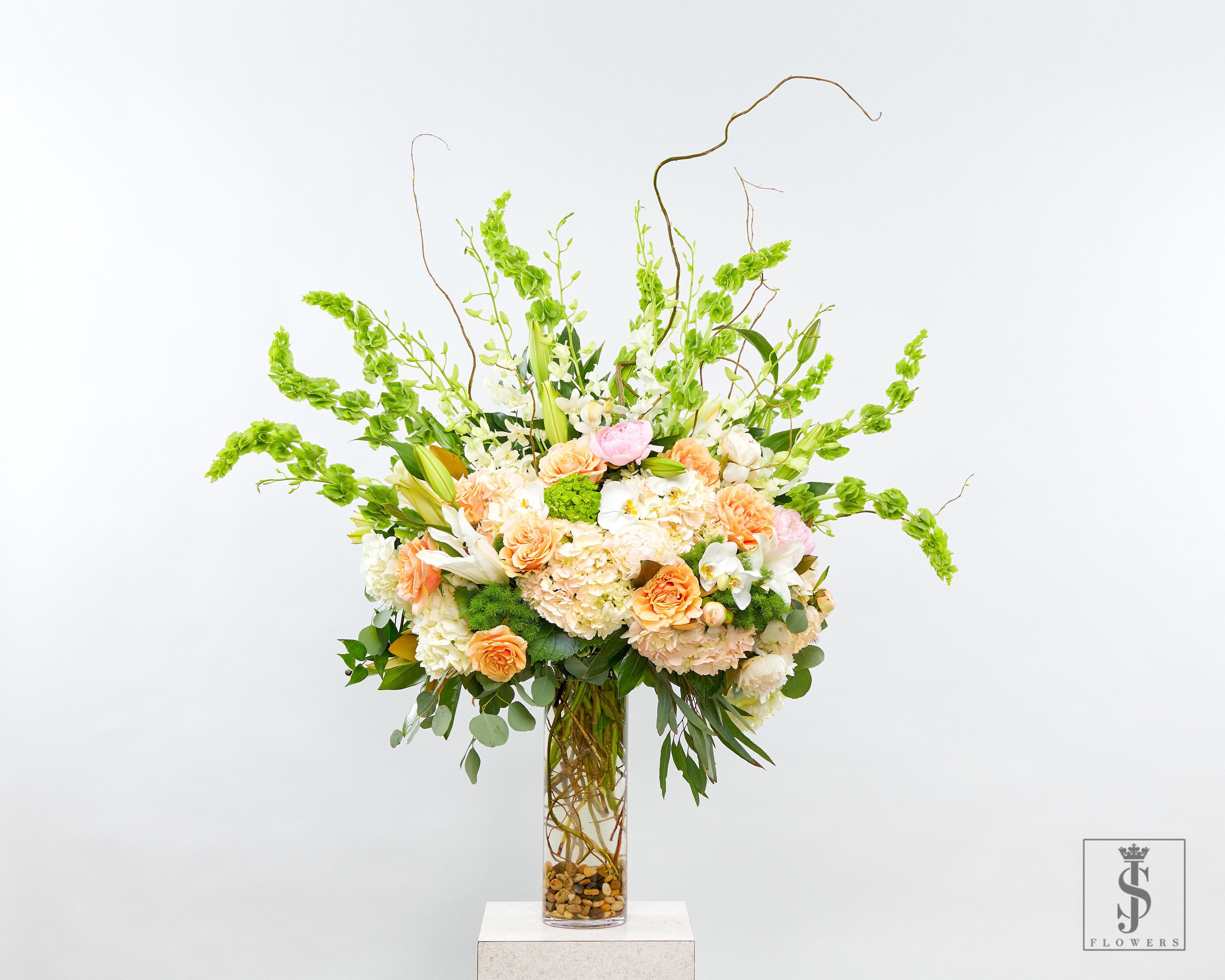 Peach Bloom Cascade - Make a bold statement with this breathtaking tall flower arrangement, designed to radiate elegance and warmth. Crafted with a luxurious mix of peach roses, white lilies, blush peonies, white hydrangeas, and exotic phalaenopsis orchids, this piece is elevated with striking Bells of Ireland and curly willow accents. The arrangement is beautifully displayed in a tall clear glass vase filled with natural stones and artistic stems, creating a stunning vertical presence perfect for grand entrances or upscale events.  Ideal for those searching for premium floral centerpieces, tall luxury flower arrangements, or elegant green and peach wedding flowers. This best-selling design is perfect for anniversaries, celebrations, sympathy arrangements, or weekly flower delivery for high-end spaces. Add timeless beauty and floral artistry to your decor with one of our most requested designer picks.  