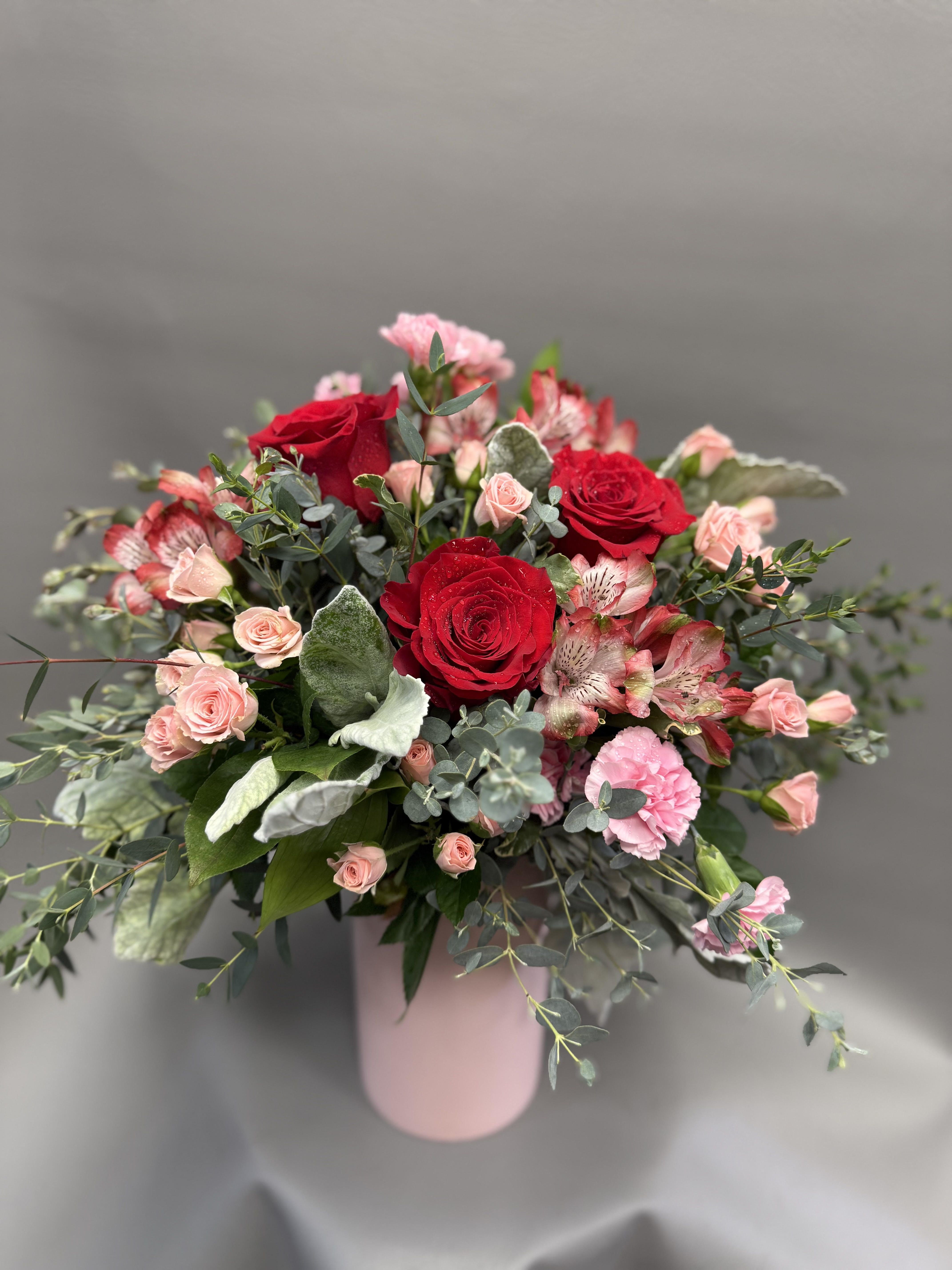 Sweetheart Rose Cylinder - Red roses meet soft blush accents in this Valentine’s favorite. Featuring red roses, blush spray roses, pink carnations, and alstroemeria with airy eucalyptus for movement and fullness. Designed in a chic pink cylinder vase—perfect for romance, anniversaries, or a heartfelt “I love you.”