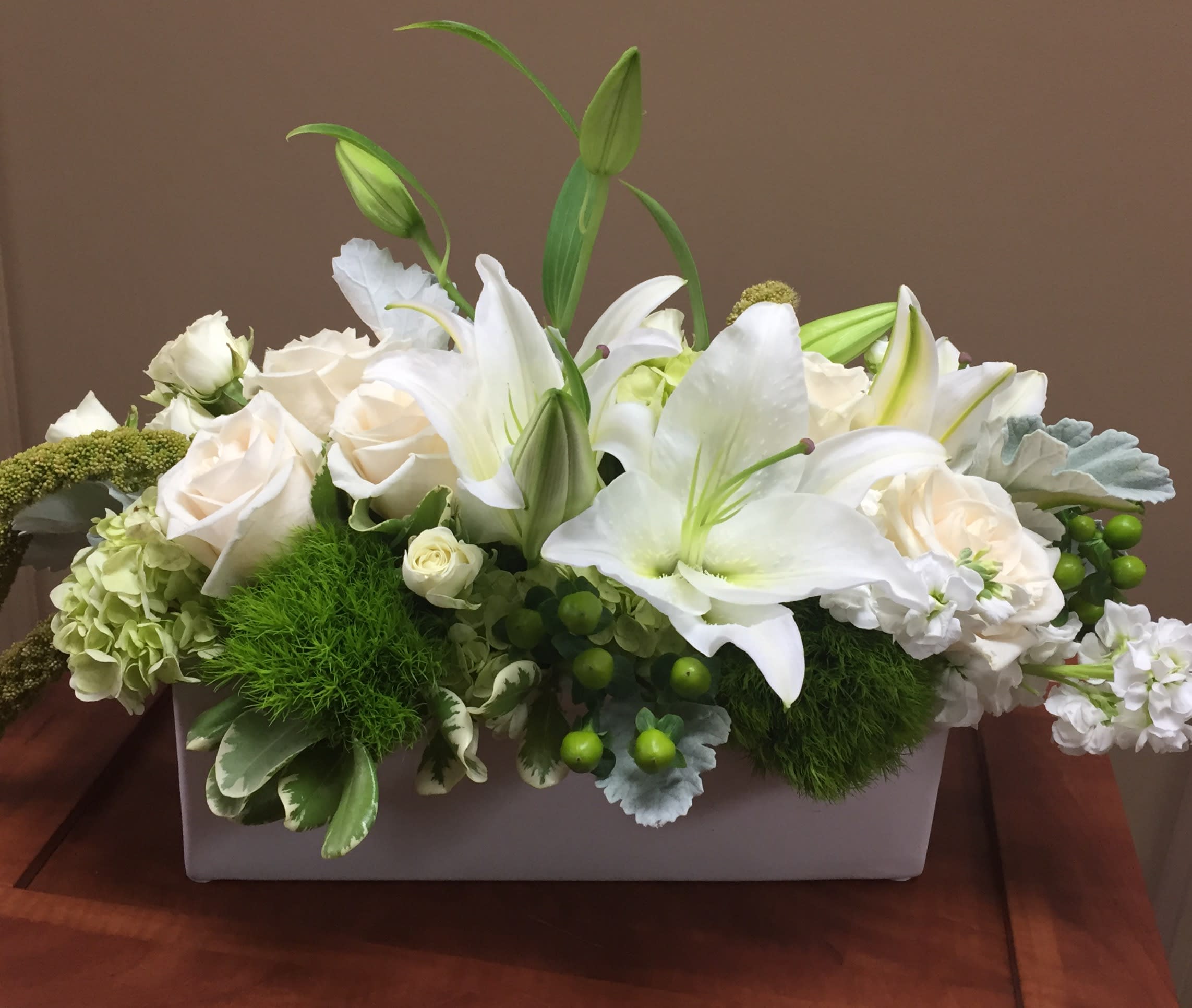 White Elegance - A beautiful and versatile design appropriate as formal dinner décor, a sympathy gift or just to let someone know they are loved. Lush with lilies, stock, roses, hydrangea and accented with textural dusty miller, trick and hypericum.