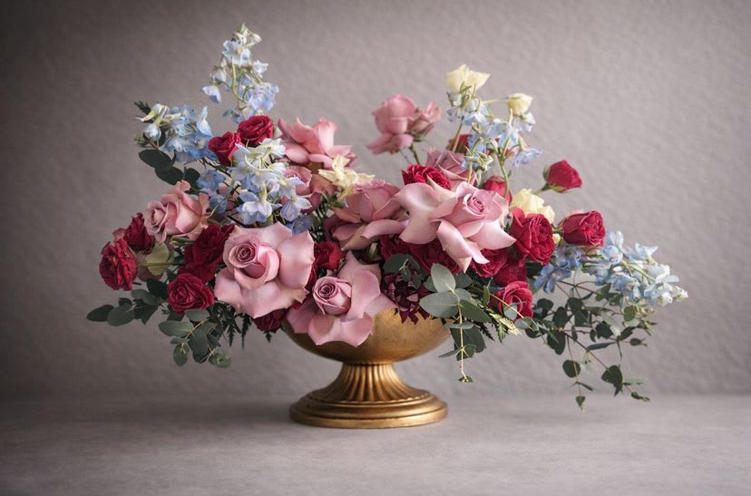 Devotion in Bloom - Devotion in Bloom is a romantic study in softness and depth—where blush and pink roses intertwine with rich crimson tones, airy blue delphinium, and cascading eucalyptus. Designed to feel intimate yet abundant, this arrangement tells a love story through layered textures and graceful movement.  Set in a classic compote, it evokes old-world romance with a modern sensibility—passionate without being loud, tender without being timid. Perfect for celebrating love in all its forms, from grand gestures to quiet, meaningful moments.