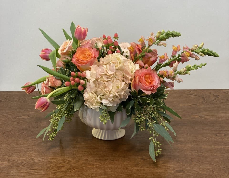  Serenade of Amour - This modern design features pink hydrangea, free spirit roses, orange snapdragons, peach spray roses, peach hypericum, and columbus tulips in a blush footed compote with a touch of seeded eucalyptus.