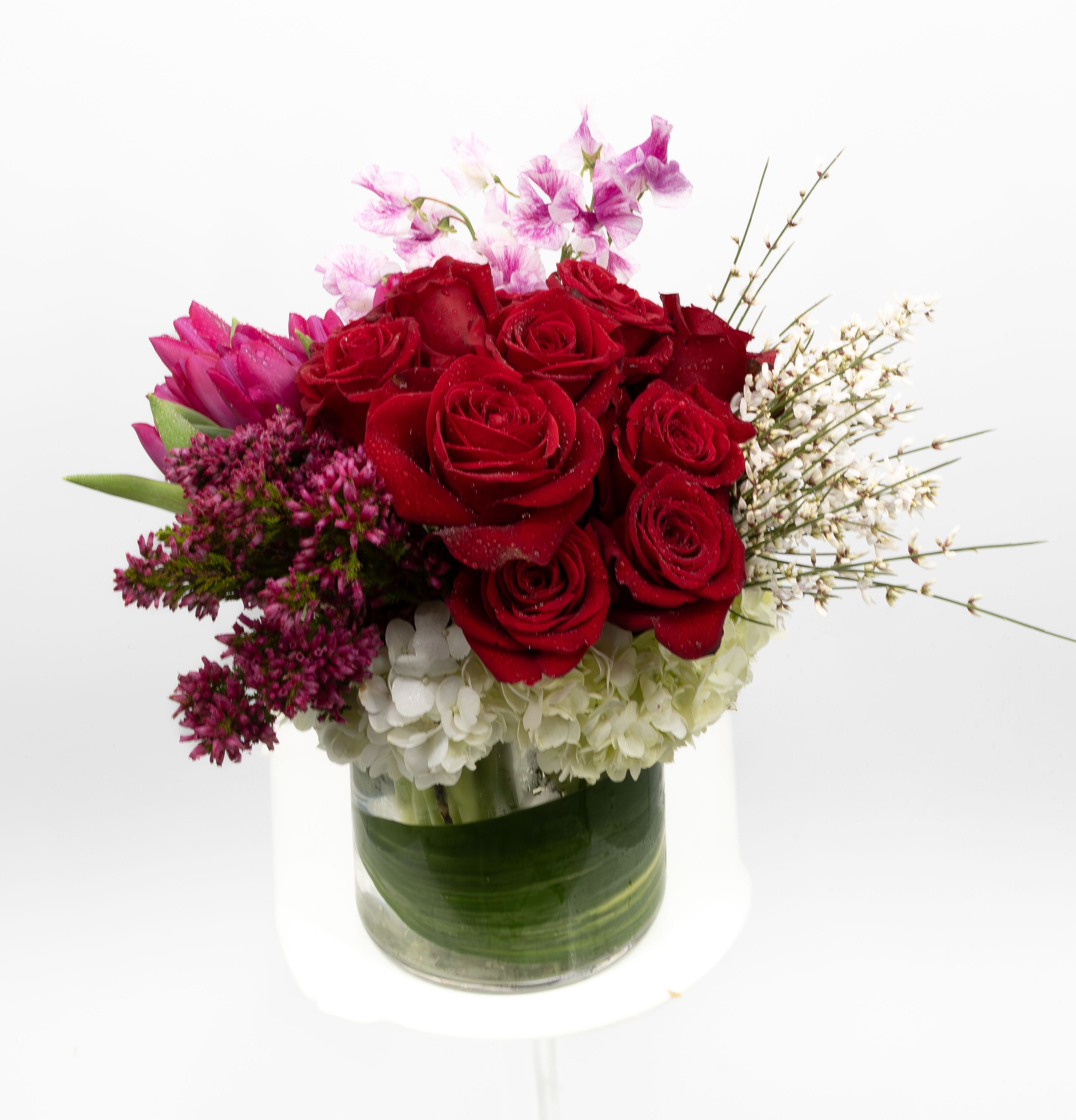 Empatîa - This arrangement showcases a charming bouquet featuring vibrant red roses, surrounded by white hydrangeas, purple flowers, and delicate pink accents, arranged beautifully in a clear cylindrical vase lined with green leaves.