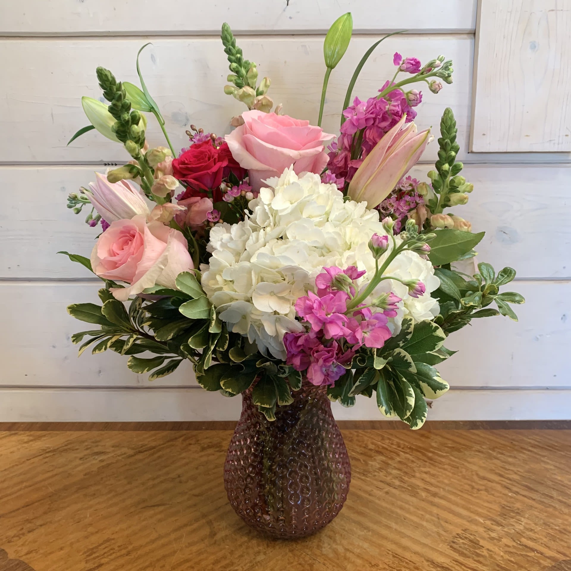Sparks Fly - White hydrangea and light pink roses, with hot pink spray roses, pink snapdragons, pink lilies, and fragrant pink stock in a pink hobnail vase with seasonal filler flower. Approximately 15" wide and 16" tall for the standard version.