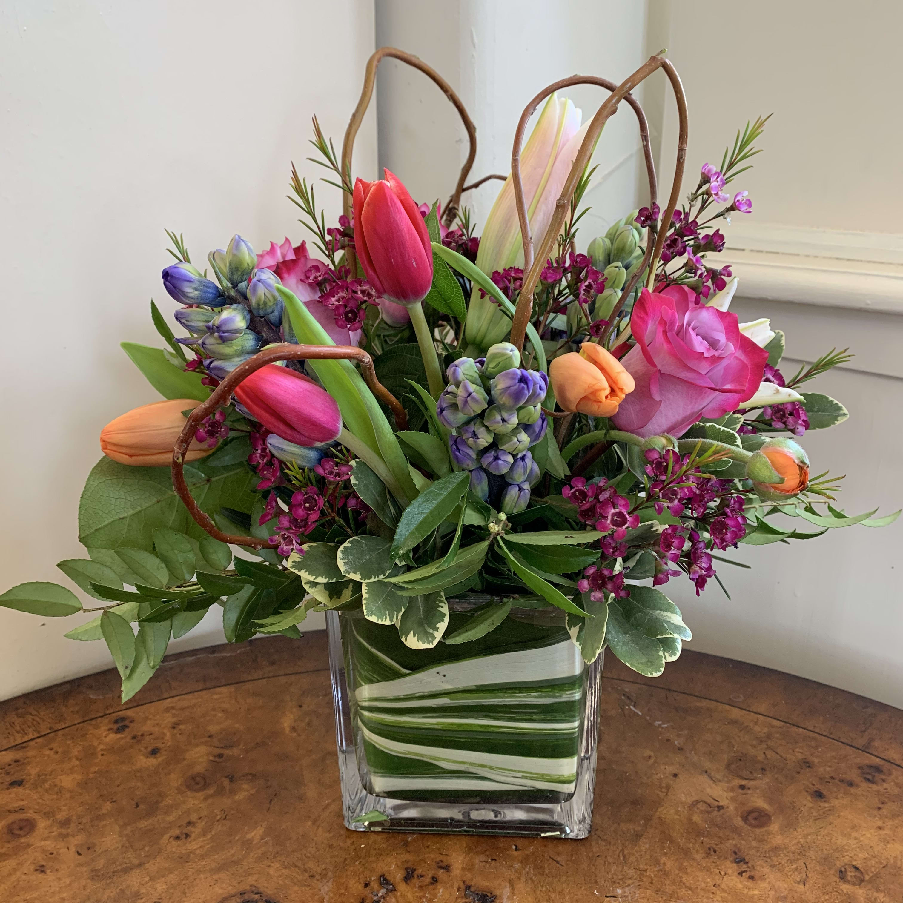 Vibrant Blooms - Lavender roses, stargazer lilies, hot pink and orange tulips, and hyacinth arranged in a leaf lined medium cube with magenta wax and curly willow loops. Approximately 12in tall and 10in wide.    