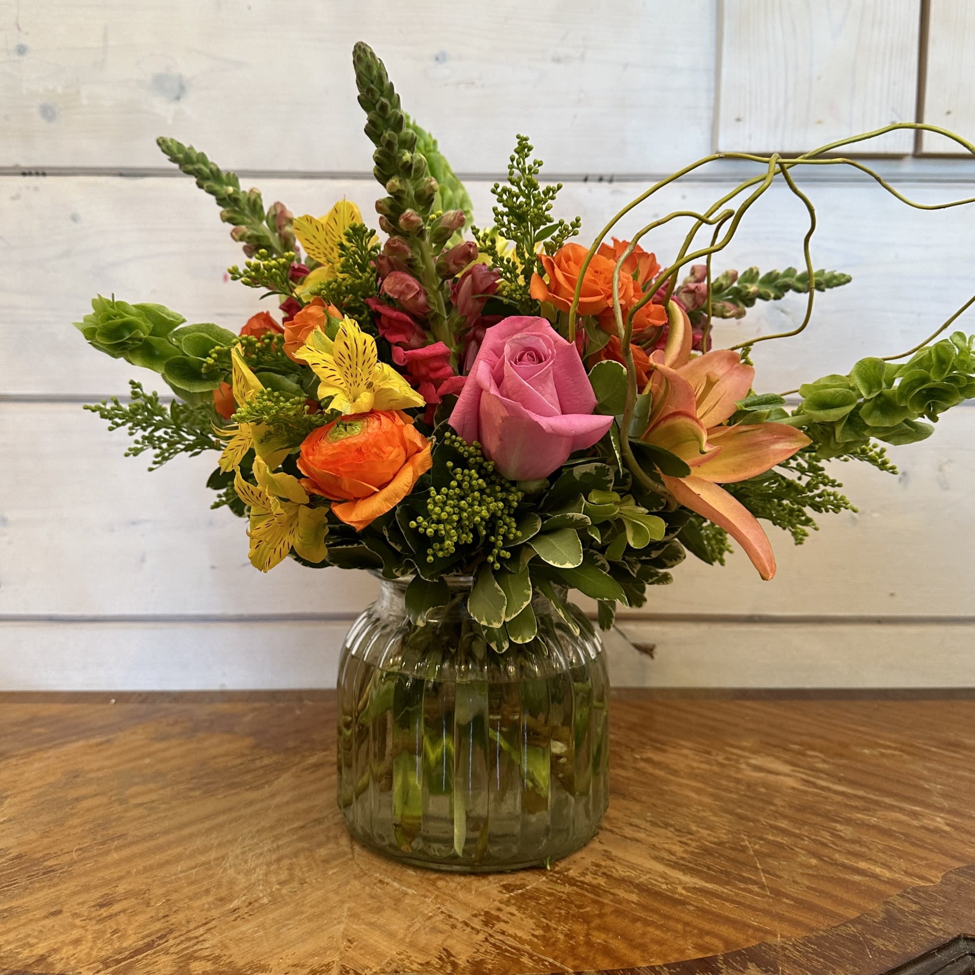 Blazing Beauty Bouquet - Pink roses, orange spray roses, bells of Ireland, hot pink snapdragons, orange lily and orange ranunculus arranged in an extra large ribbed glass ginger jar with solidago, yellow alstroemeria and looped curly willow. Approximately 14in tall and 13in wide. 