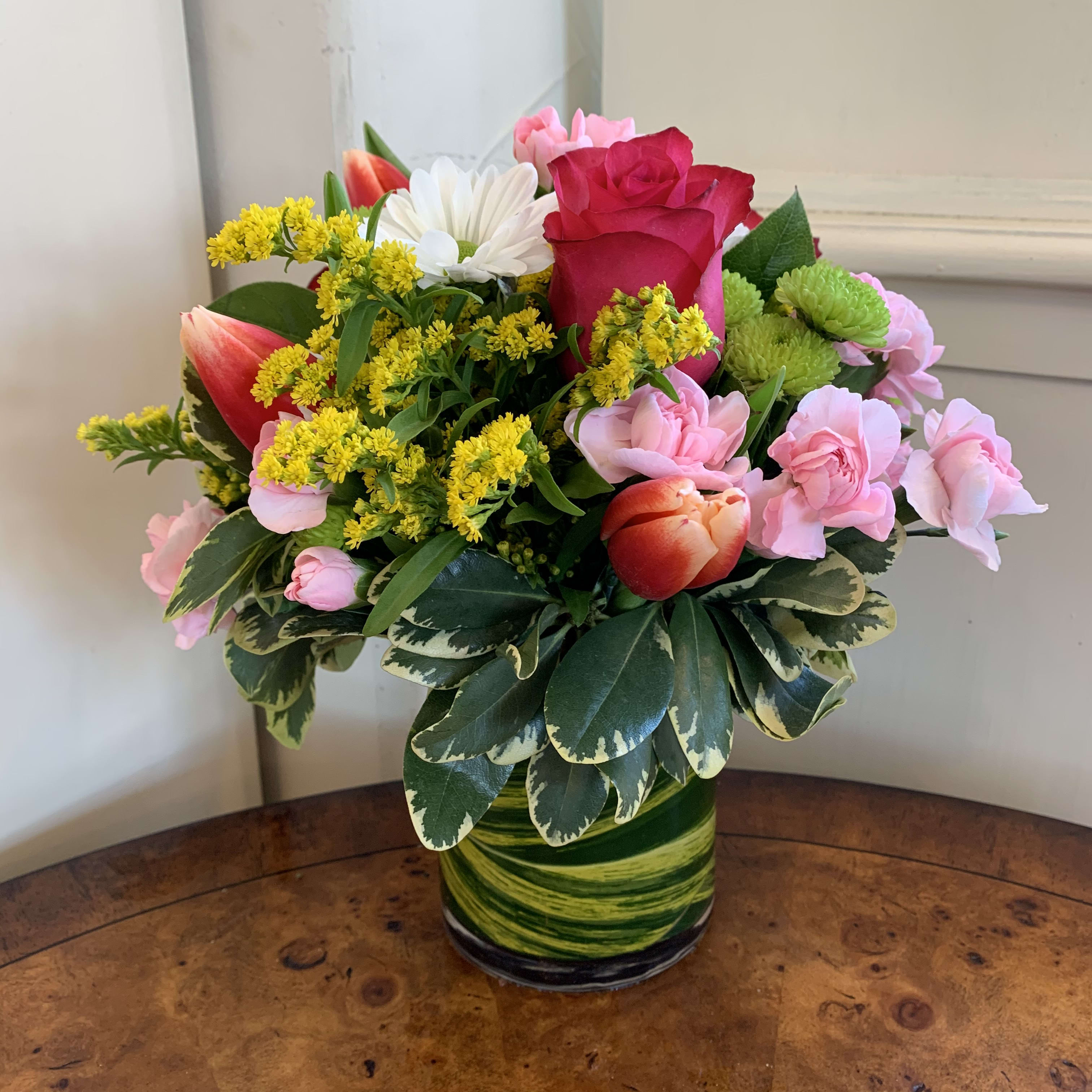 Tulip's Kiss  - Hot pink roses, orange or pink tulips, green buttons, white daisies, and pink mini carnations arranged in a small leaf lined gather vase with solidago or other complimentary filler flowers. Approximately 8in tall and 8 in wide. 