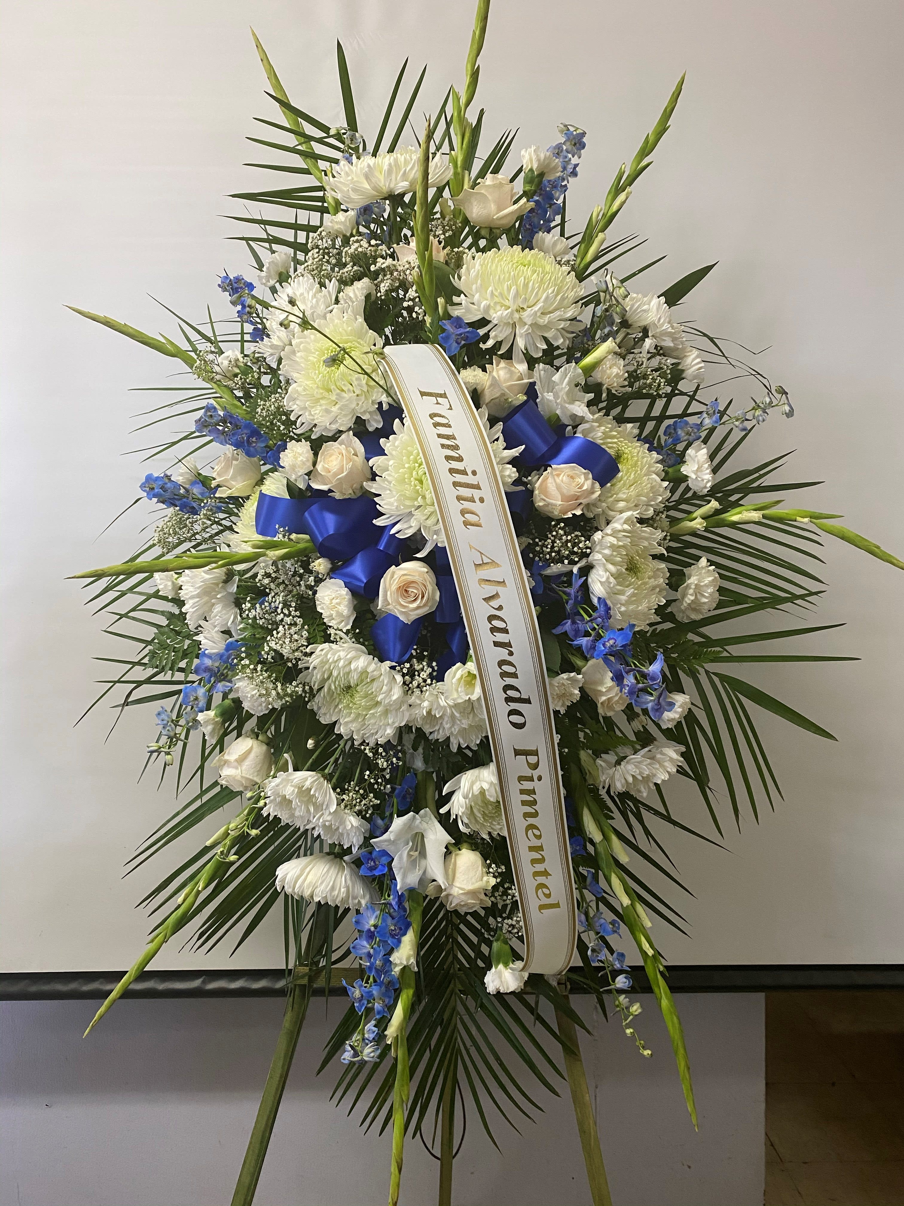 Sympathy Standing Spray #39 - Standing spray of white fresh cut flowers that includes a touch of blue. Embellished with blue ribbon loops and bow. Colors can be modified depending on availability. Designed on an easel which is included with arrangement and does NOT need to be returned. Banner is included with arrangement. Please specify what is to be printed on banner upon check out under "FLORIST INSTRUCTIONS". LIMITED TO 5 WORDS ON BANNER.