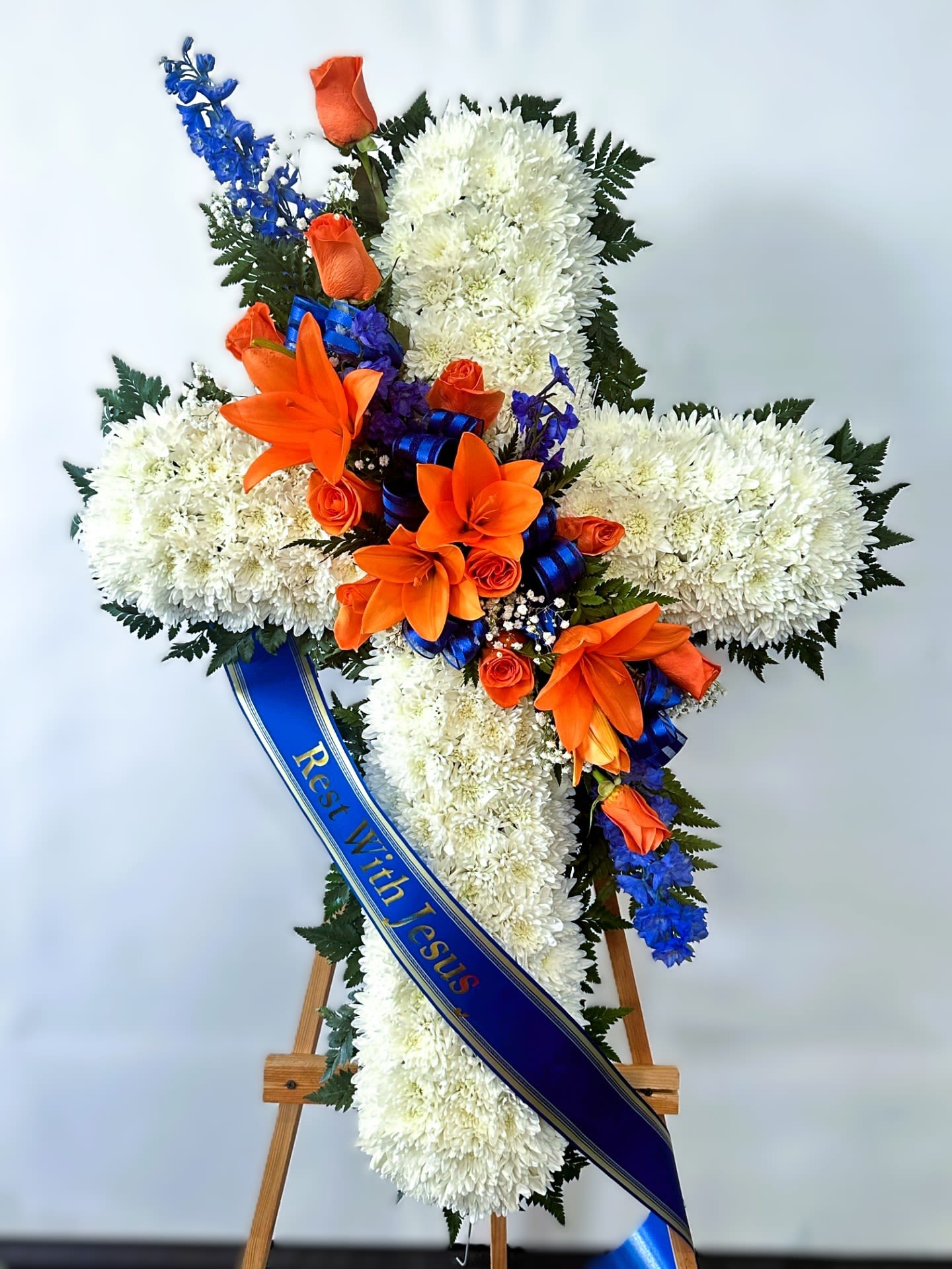 Sympathy Cross #45 - All white standing cross with an orange and blue cluster of fresh cut flowers. Colors can be modified depending on availability. Designed on an easel that is included with the arrangement and does NOT need to be returned. Banner is included with arrangement. Please specify what is to be printed on banner upon check out under "FLORIST INSTRUCTIONS". LIMITED TO 5 WORDS ON BANNER.