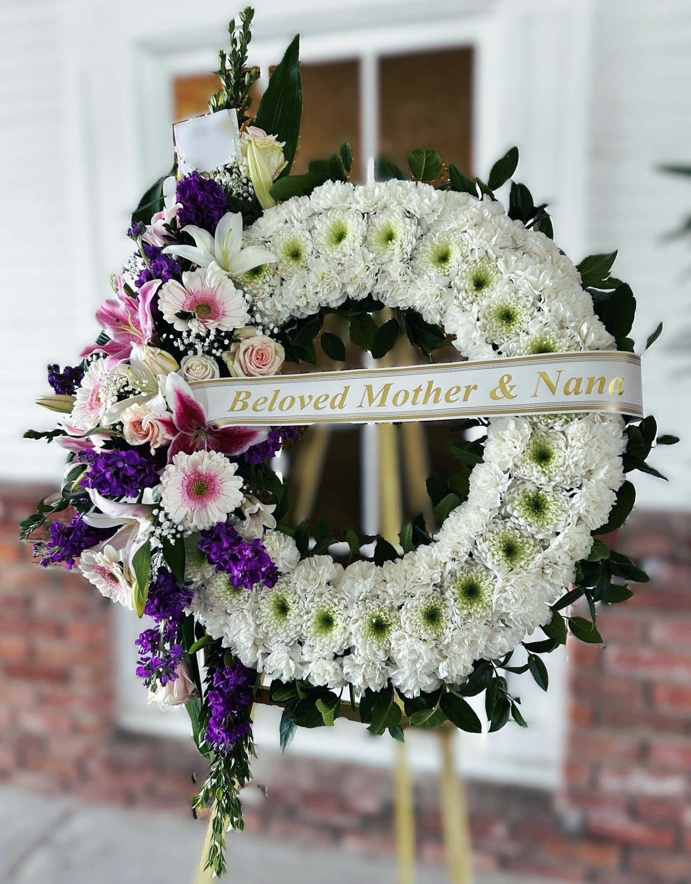 Sympathy Wreath #50 - All white standing wreath with pink and purple cluster made of fresh cut flowers. Colors can be modified depending on availability. Designed on an easel that is included with the arrangement and does NOT need to be returned. Banner is included with arrangement. Please specify what is to be printed on banner upon check out under "FLORIST INSTRUCTIONS". LIMITED TO 5 WORDS ON BANNER. If no note is added as to what to put NO banner will be added.