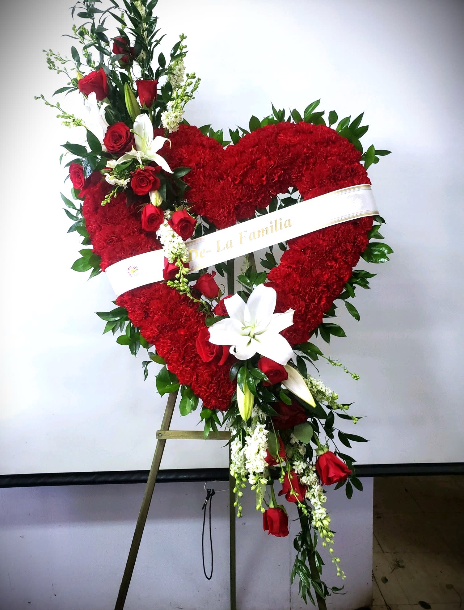 Sympathy Open Heart #63 - Standing open heart made of all red carnations embellished with a a cluster of red roses and white lilies. Colors can be modified depending on availability. Designed on an easel that is included with the arrangement and does NOT need to be returned. Banner NOT included.