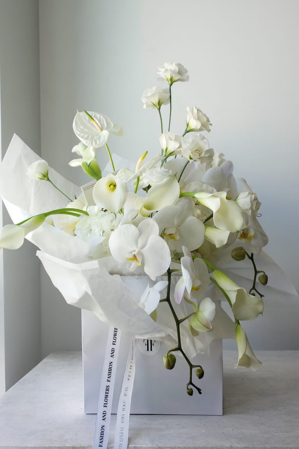 White Couture - White Couture is a refined all-white arrangement designed in our signature Fashion &amp; Flowers aesthetic. Sculptural phalaenopsis orchids, elegant calla lilies, lisianthus and anthuriums create a clean, architectural silhouette with a soft, luxurious flow. This piece embodies quiet luxury and timeless elegance — perfect for celebrations, refined gifting or elevating any interior.  The arrangement is styled in a white tall ceramic vase, adding height, elegance and a modern gallery-like feel to the composition.