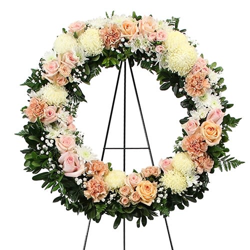 Heavenly Halo - A timeless sympathy wreath designed in soft, comforting tones of blush, peach, and ivory. Featuring roses and spray roses, white cushion mums, peach carnations, and delicate baby’s breath, finished with lush greenery in a full round shape. A beautiful tribute to share love, peace, and heartfelt condolences. 22 inches