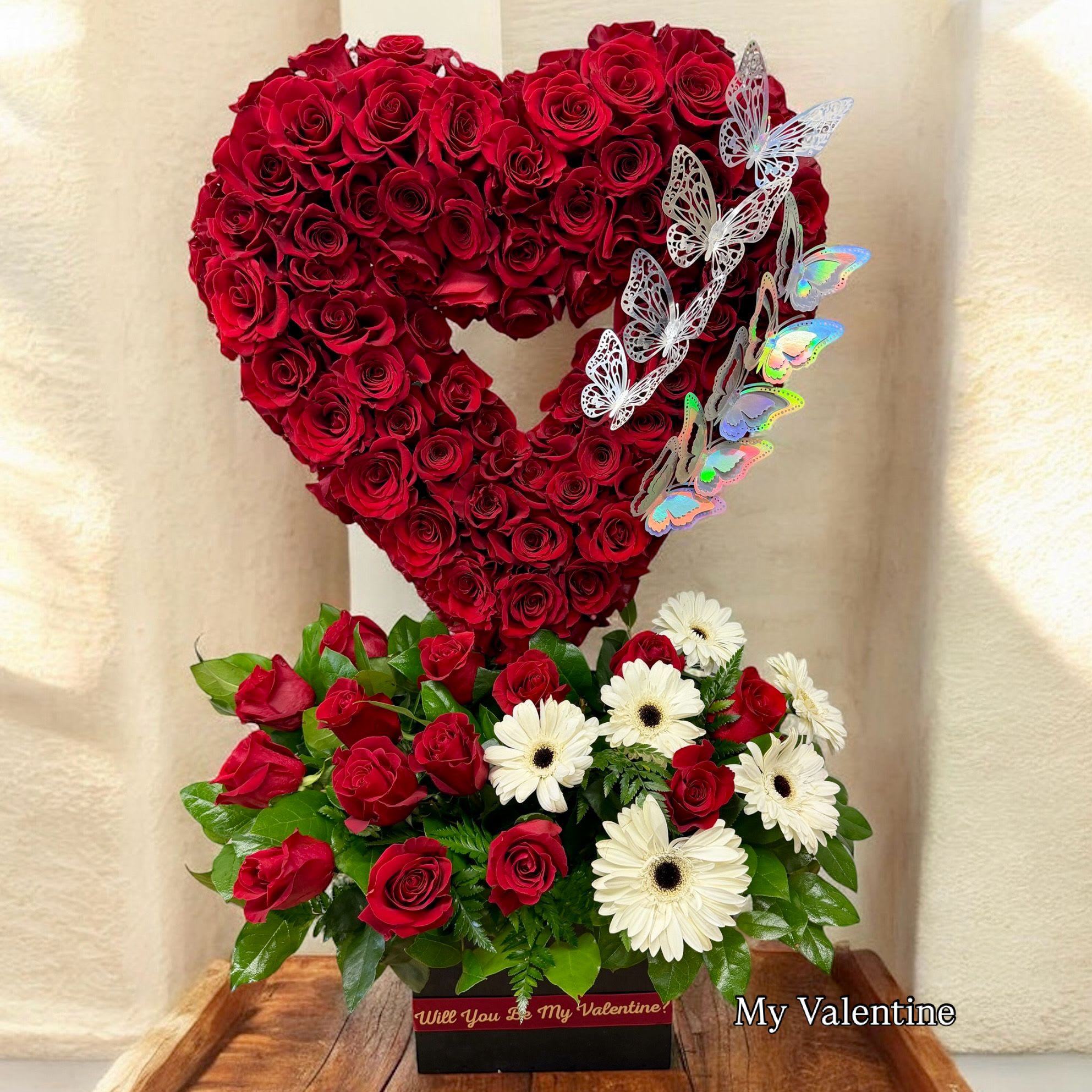 My Valentine - A bold and romantic Valentine’s arrangement featuring a stunning open-heart design made with deep red roses, accented with elegant butterfly details. Below, a lush garden-style base mixes red roses with crisp white gerbera daisies and fresh greenery, all presented in a keepsake box that reads “Will You Be My Valentine?” — a perfect statement piece for love and romance.