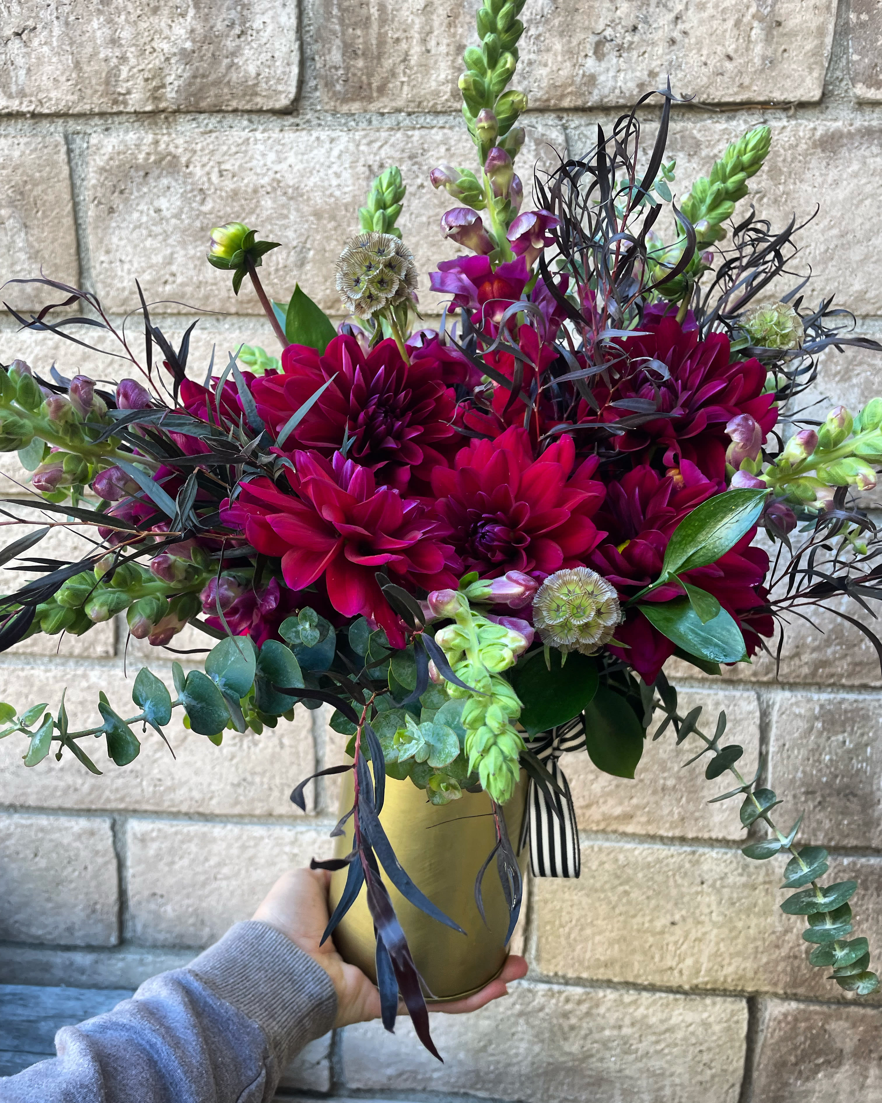 The Plum Dahlia - Here she is, a lush and bold arrangement of our signature flower- The Plum Dahlia. A little bit burgundy, a little bit purple, a lot of drama in a tall gold vase. Mixed with fragrant eucalyptus and burgundy Snapdragons, this is a distinct gift for a lover of Dahlias and rich color. 