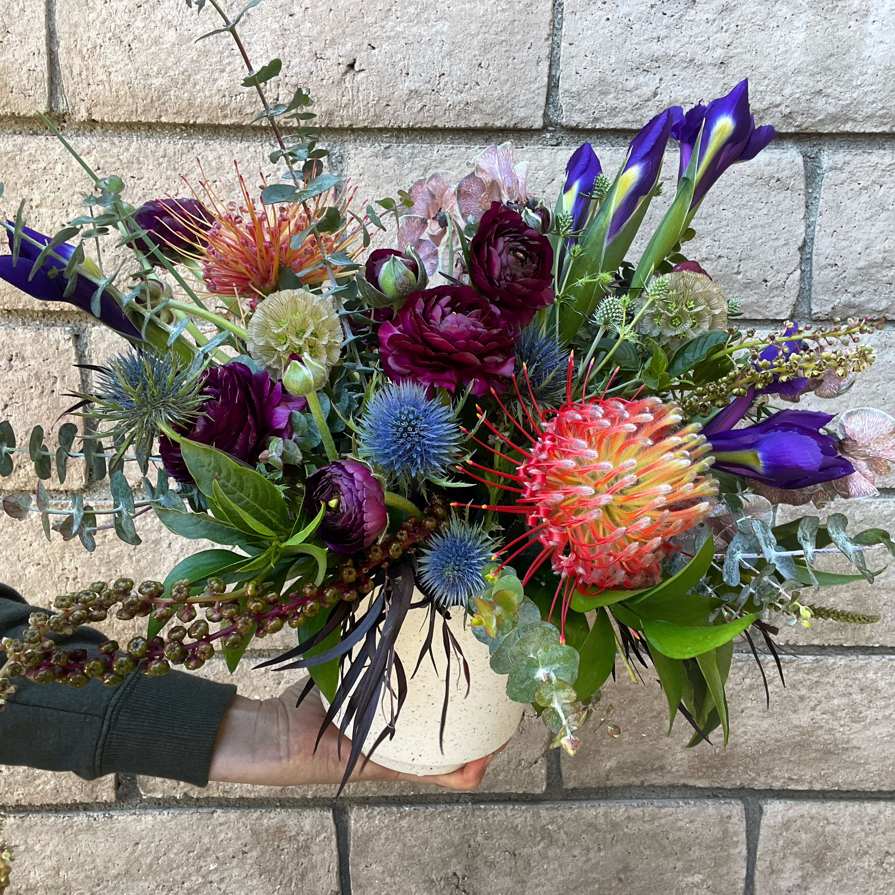 Wild Child - For the lover of everything wild and weird and wonderful. A raucous mix of long lasting and highly textured botanicals and flowers, including Pincushion Protea, purple Iris, burgundy Ranunculus, blue Thistle and Scabiosa pods in a fragrant bed of Eucalyptus. Every design will vary just a bit, but the spirit and tone of this design will shine through. 