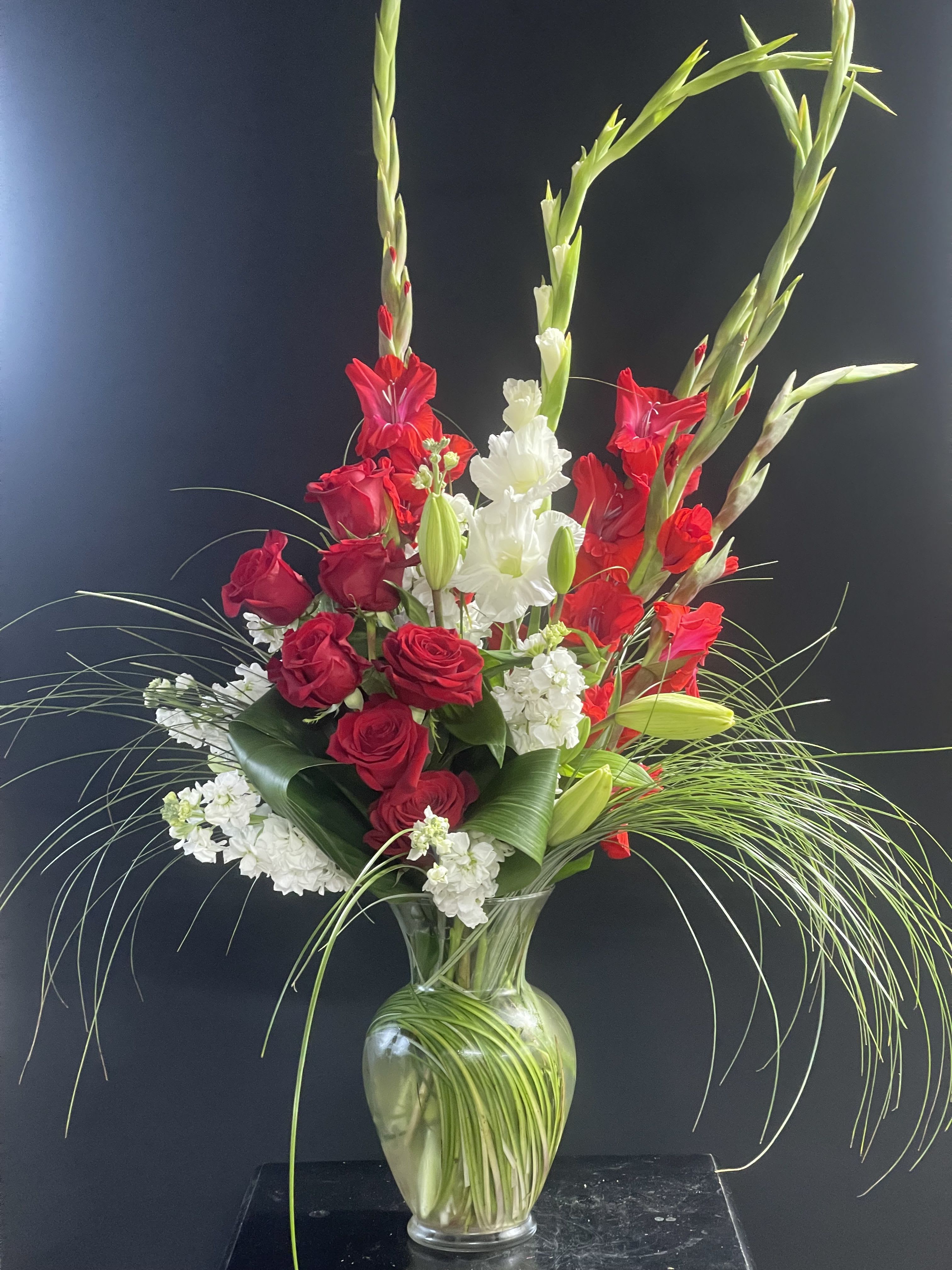 Ivory and Flame Floral Arrangement - A striking floral composition that marries the bold passion of red and white gladiolus and white lilies with radiant red roses nestled in a bouquet of aspidistra leaves, white hydrangeas, and stock. Accented with whispering grasses, this vased arrangement brings dramatic contrast and graceful balance — perfect for commanding attention at any grand event. Ivory and Flame makes a bold statement.
