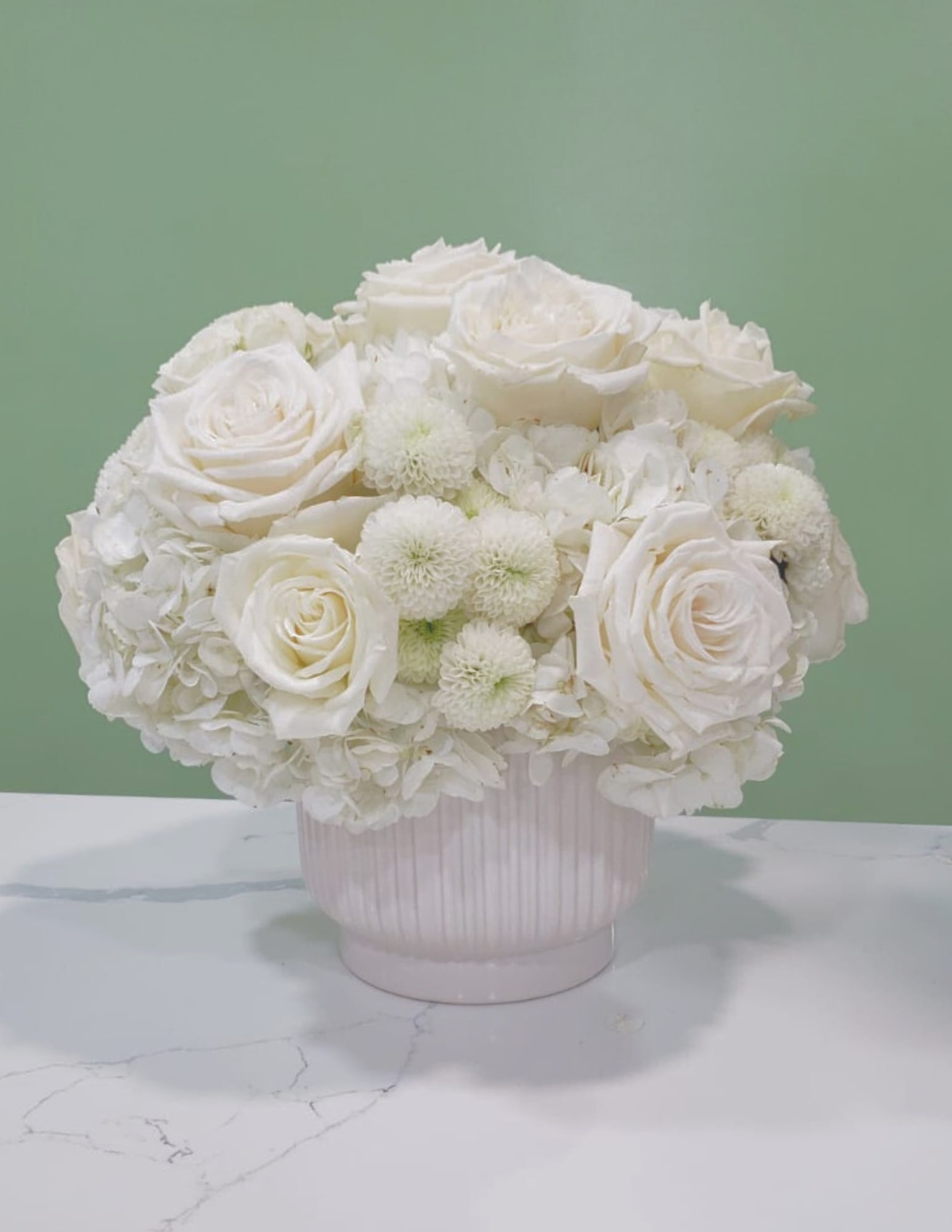 Magnifique - This all white arrangement has hydrangeas, roses, and pompoms in a white ceramic vase.