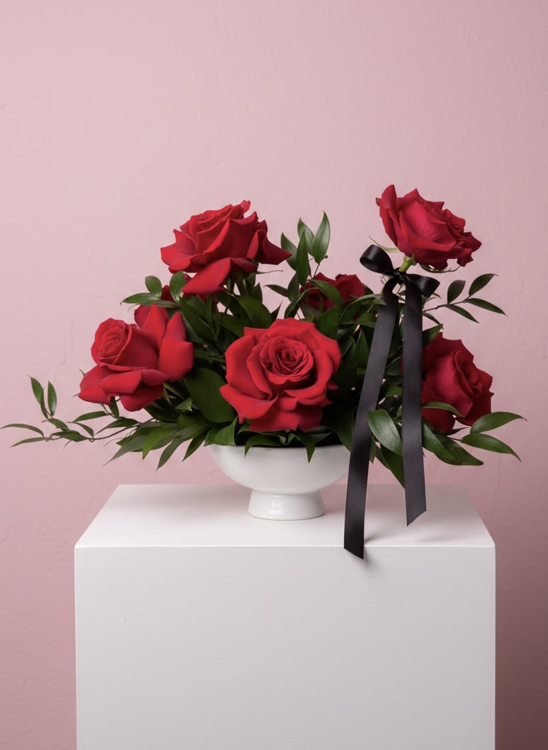 Cupids Arrow - Classic red roses with fresh greenery, accented with a black ribbon and arranged in a timeless white pedestal vase.
