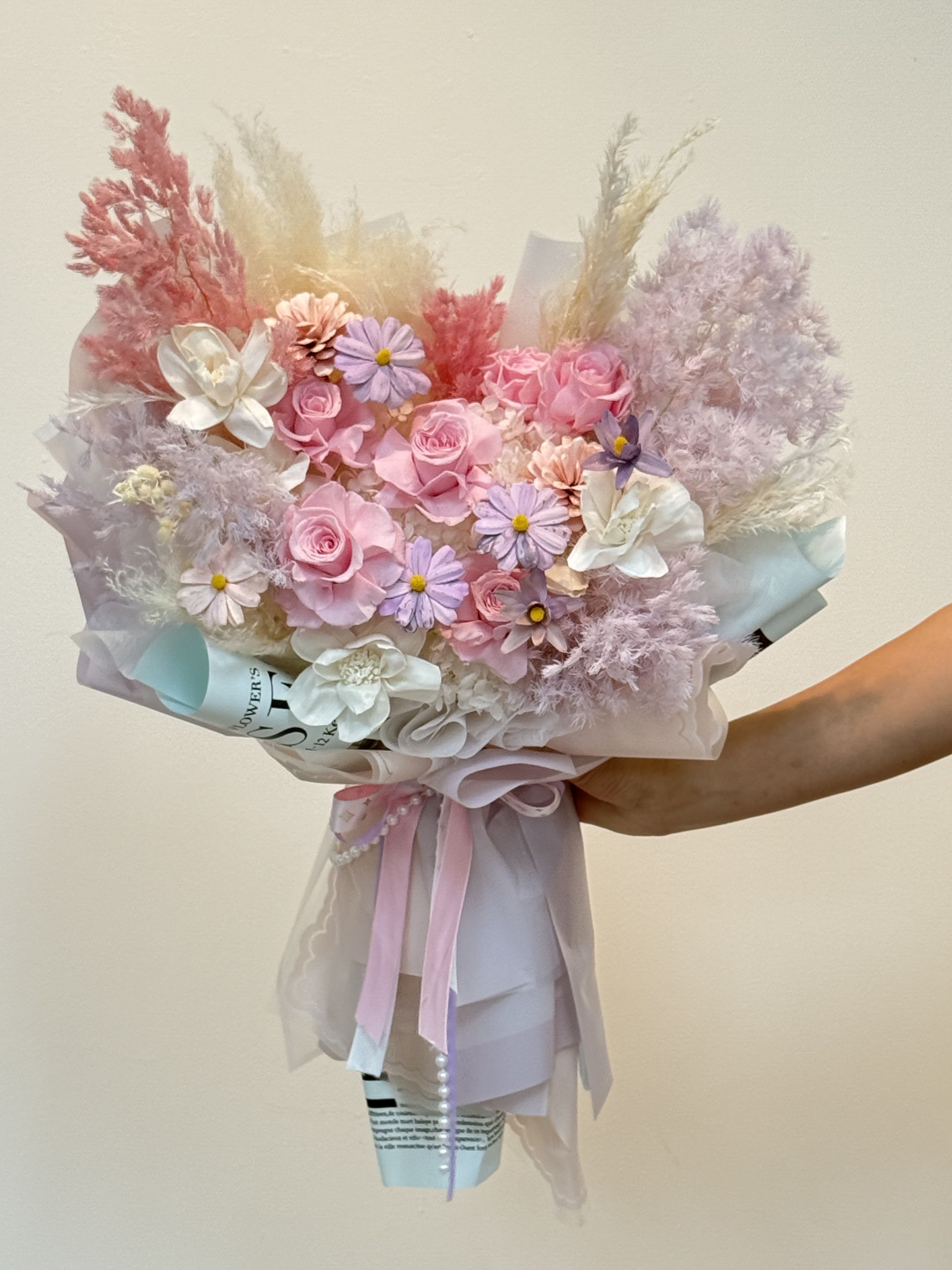 Love in Pastel - This is a mixed bouquet of soft pink and white. It adopts a variety of floral materials and special packaging designs, with a sweet, dreamy and elegant style. Composition of flower materials Roses: Light pink and white roses are the main focal flowers. Matching flowers: including white or light purple orange stalks, small daisies and other filling flowers. Feathers/hay: pink and beige rabbittail grass, reeds or feather-like decorations add a soft and elegant texture. Leaf material: dyed light blue or gray-green eucalyptus leaves are used as embellishments to provide color contrast. Packaging style The bouquet adopts Korean packaging style and is wrapped in multiple layers of light pink, white and light blue wrapping paper or organza. This multi-layered, soft-toned packaging highlights the dreamy feeling of flowers. Finally, it was tied with a light pink ribbon and decorated with pearl jewelry. Symbolic meaning This bouquet mainly symbolizes gentleness, purity, romance and eternal love. The use of dried flowers and preserved flowers means that this beauty can be preserved for a long time. It is very suitable for birthday gifts, anniversary gifts, or occasions to express tenderness.