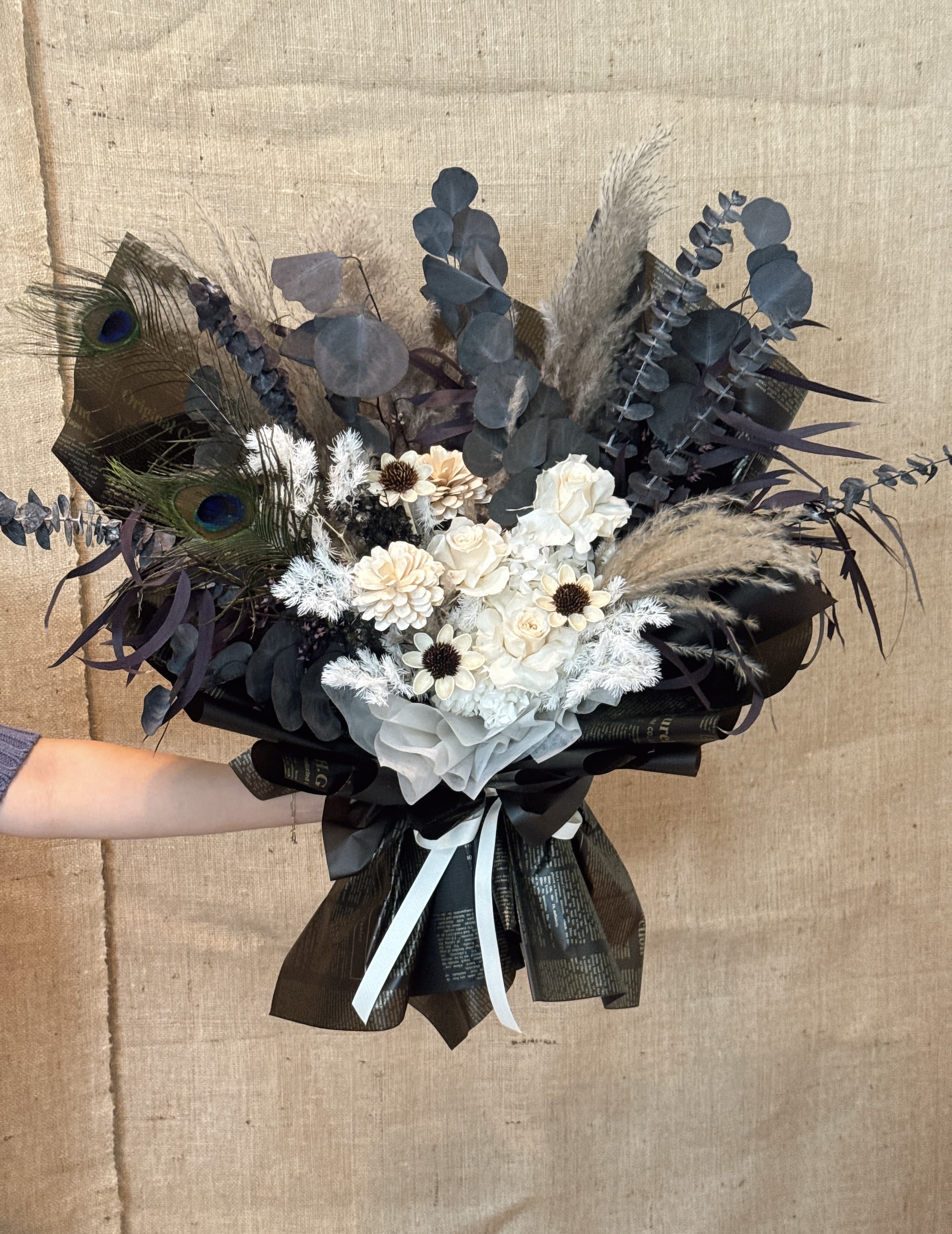 Eternal Noir - This is an exquisite black and white bouquet, which is popular for its unique color matching and symbolism. Color The main body of the bouquet is black and white flowers of varying shades, supplemented by dark leaves and beige apricot or light-colored wrapping paper. The black packaging can set off the beauty of the flower itself. Flower materials The main flower materials may include white roses, beige small flowers, white hydrangeas and dark leaves of eucalytus leaves, etc. Black and white bouquets usually also symbolize reason, peace and purity. At the same time, the peacock feathers added to the bouquet imply auspiciousness, official power and wealth and luxury in Chinese culture. Occasion This bouquet is very suitable as a birthday gift, Valentine's Day gift, marriage proposal bouquet or any anniversary gift that needs to express unique and sincere feelings.