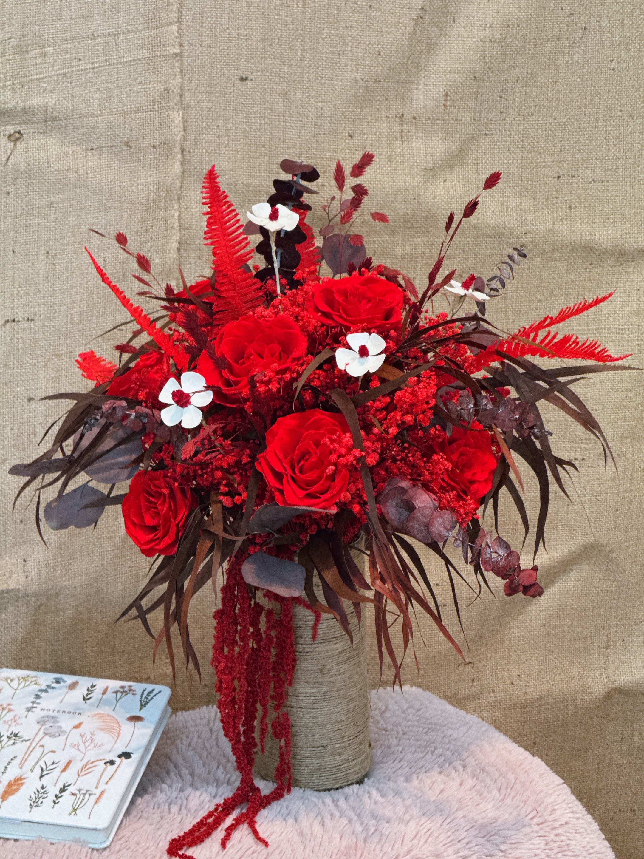 Timeless Bloom - This vase flower arrangement is mainly made of bright red roses, with crimson filling flowers and small white decorative flowers, creating a strong visual contrast. In flower arrangement, dark brown leaves and red ferns are also used as leaves to add a sense of layering and texture. The vase adopts a cylindrical container wrapped with coarse twine, so that the overall style has a natural and simple texture. The long red streamer hangs from the bouquet, enhancing the sense of dynamism and decoration. The overall design is warm in color, full of drama and modernity, suitable for festivals or special celebrations.
