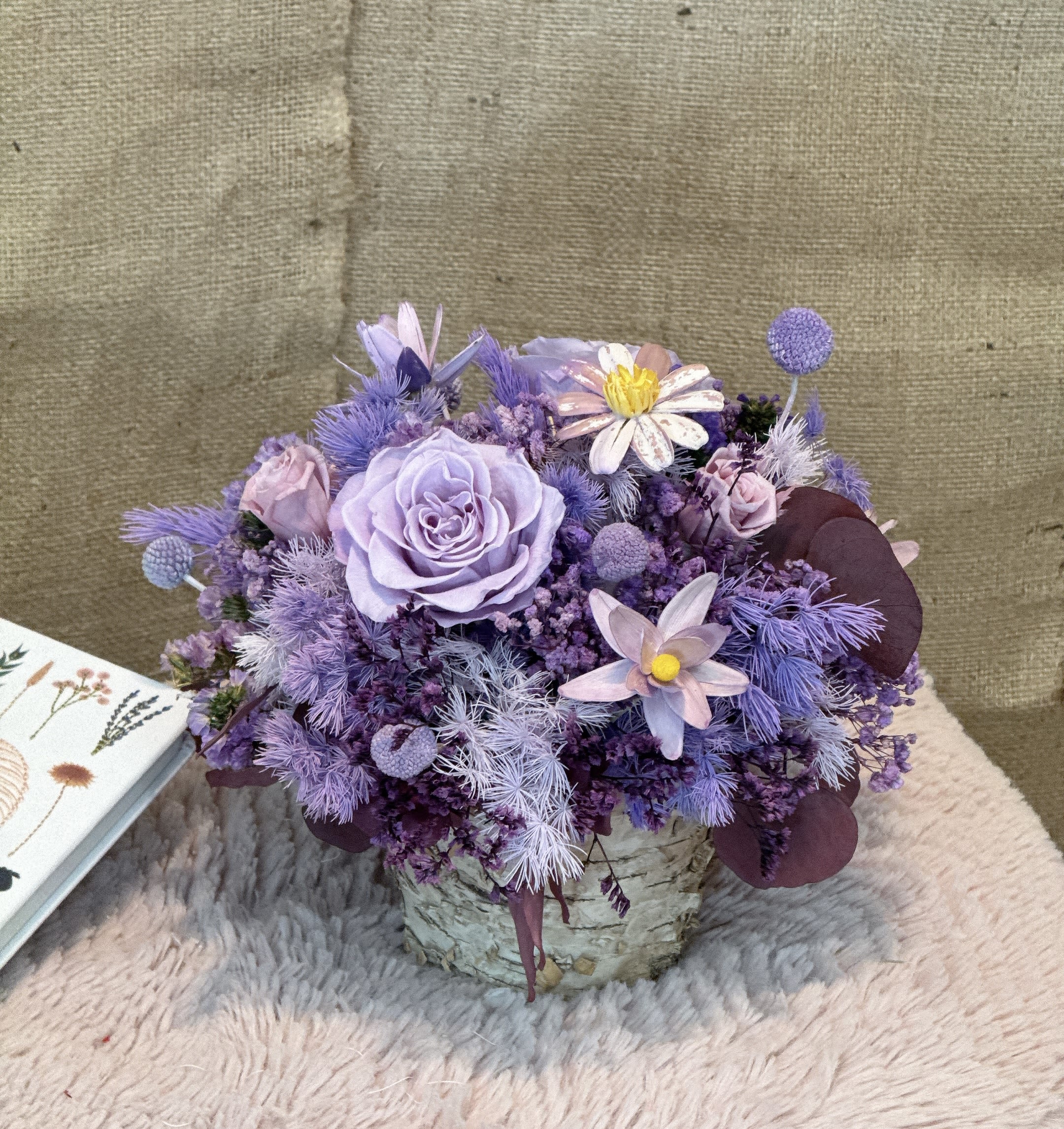 Lavender Still - This is a very exquisite combination of purple flowers, including a variety of flower materials and decorations. The main features include: 1. *Main flower material*: The center is a large purple rose (or similar purple flower), which becomes the visual focus. 2. *Auxiliary flower materials*: with purple small pompoms (similar to thistles), white daisies with yellow centers, and purple and white decorative flowers. 3. *Filling material*: Purple flower branches and white feather-like decorations are used to make the whole fuller. 4. *Container*: The bouquet is arranged in a ceramic or glass flower pot with a white texture, adding texture. 5. *Color matching*: The overall color is purple, with white and dark purple leaves, presenting an elegant and layered visual effect. This bouquet of flowers is suitable for decoration, gifts or special occasions, showing exquisite color matching and floral design.