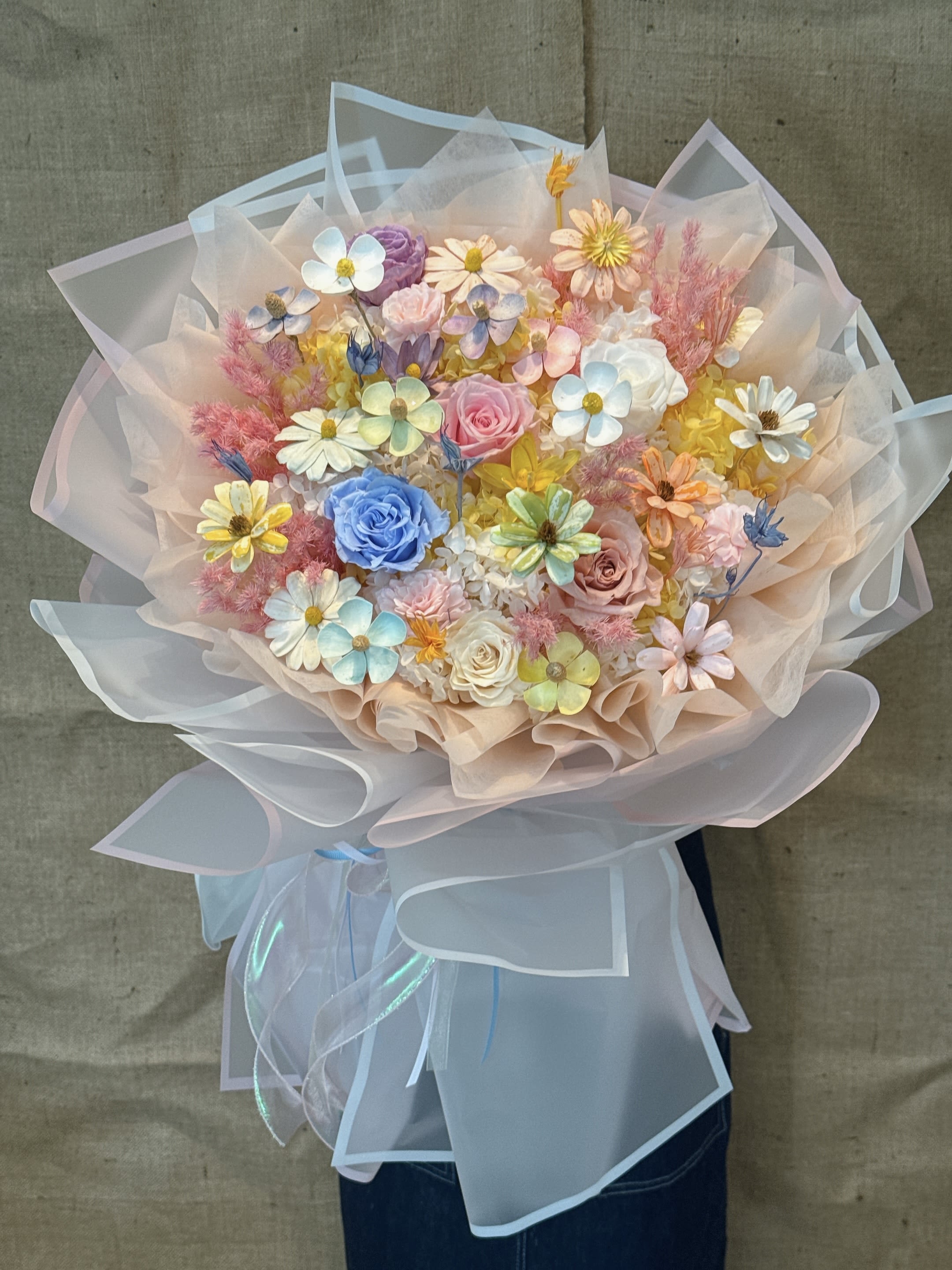 Colorful Collection - This is a colorful mixed bouquet. This bouquet of flowers combines a variety of bright colors and different types of flowers, presenting a lively, bright and energetic style. Flower materials and color matching The bouquet adopts a bold and harmonious color combination, including: Blue rose: an eye-catching blue flower in the center, symbolizing mystery, miracle or unique love. Yellow daisies: Many small yellow flowers dot it, bringing a sense of sunshine and hope. Pink and white flowers: mix and match pink, white and cream rose-like flowers and small flowers to create a soft and romantic atmosphere. Other materials: with pink feather-like or grass-like decorations to increase the texture and layering. Style and packaging The bouquet adopts the popular Korean ins style packaging: Wrapping paper: White and transparent organza or tulle are wrapped in layers to create a light, dreamy and fluffy effect. Design: The overall design focuses on color matching and photo effect, with high visual attractiveness, suitable for people who pursue fashion and personality. Meaning and use Meaning: This bouquet is rich in meaning. Blue roses represent impossible love or unique charm. Small yellow flowers bring joy and convey positive, colorful and special blessings as a whole. Occasion: Suitable for birthday gifts, graduation gifts, holiday surprises or any cheerful occasions worth celebrating